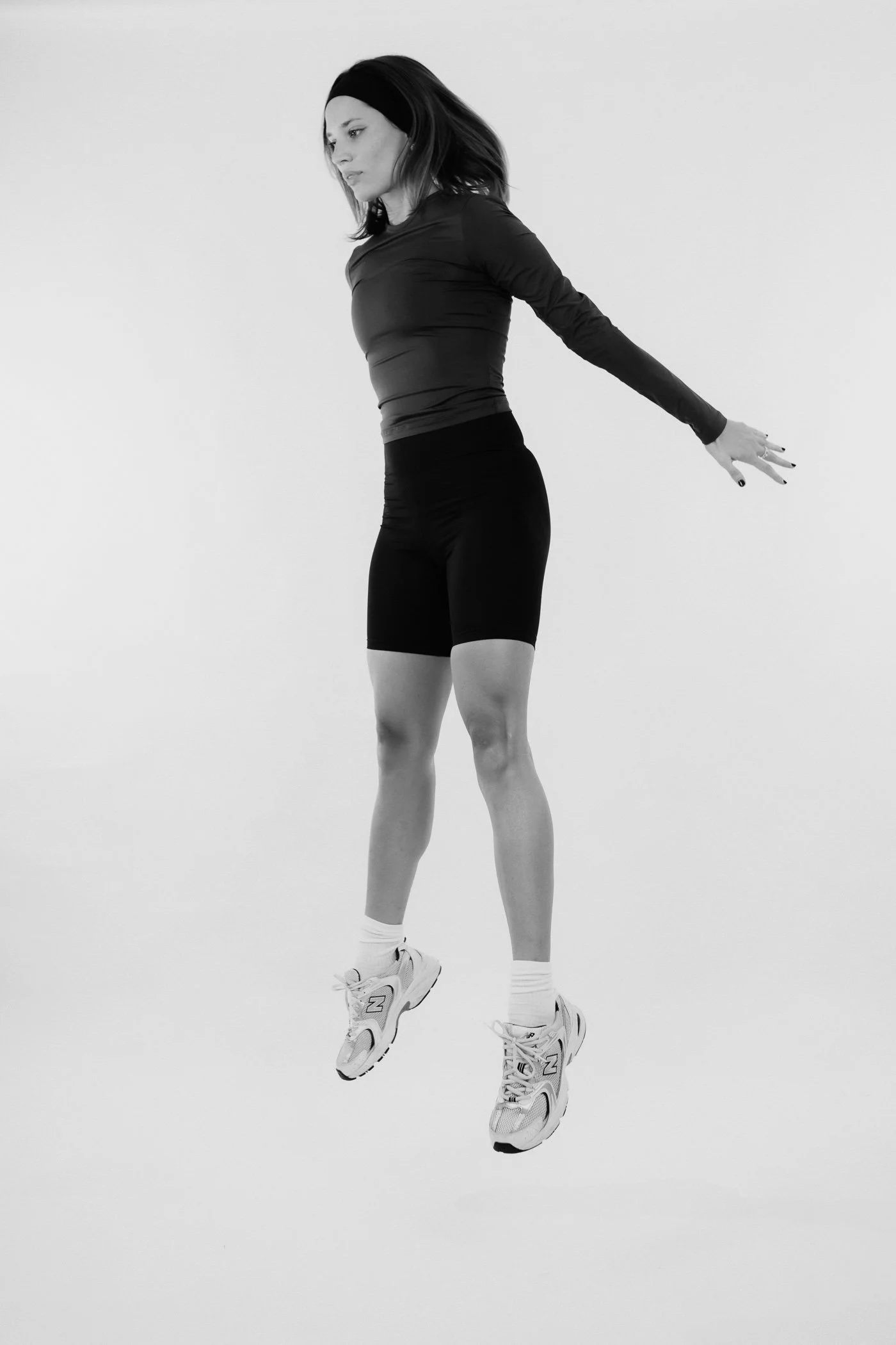Donna che salta in aria con i piedi rivolti verso il basso, indossa scarpe da ginnastica bianche e abbigliamento sportivo nero, con sfondo bianco.