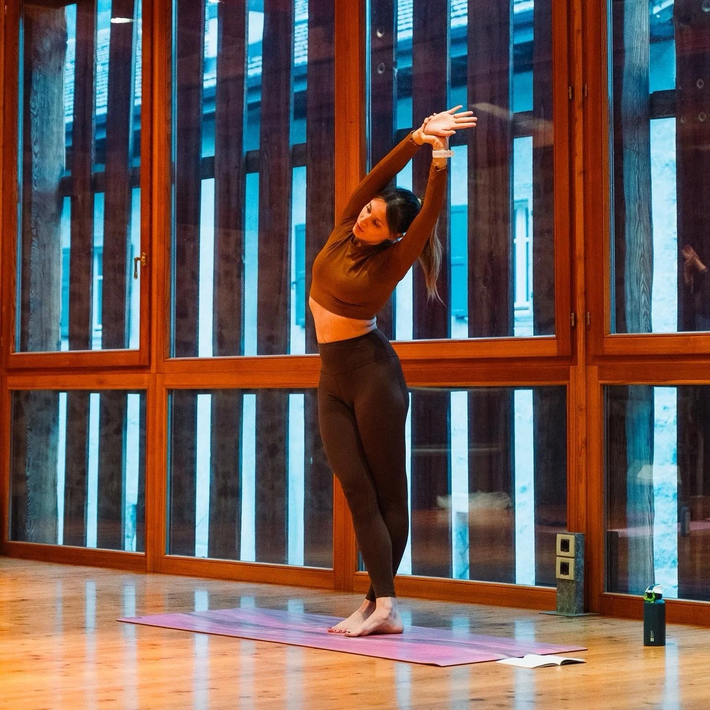 Una donna che fa esercizi di stretching su un tappetino in una stanza con grandi finestre di legno, indossando abbigliamento sportivo marrone e leggings neri.