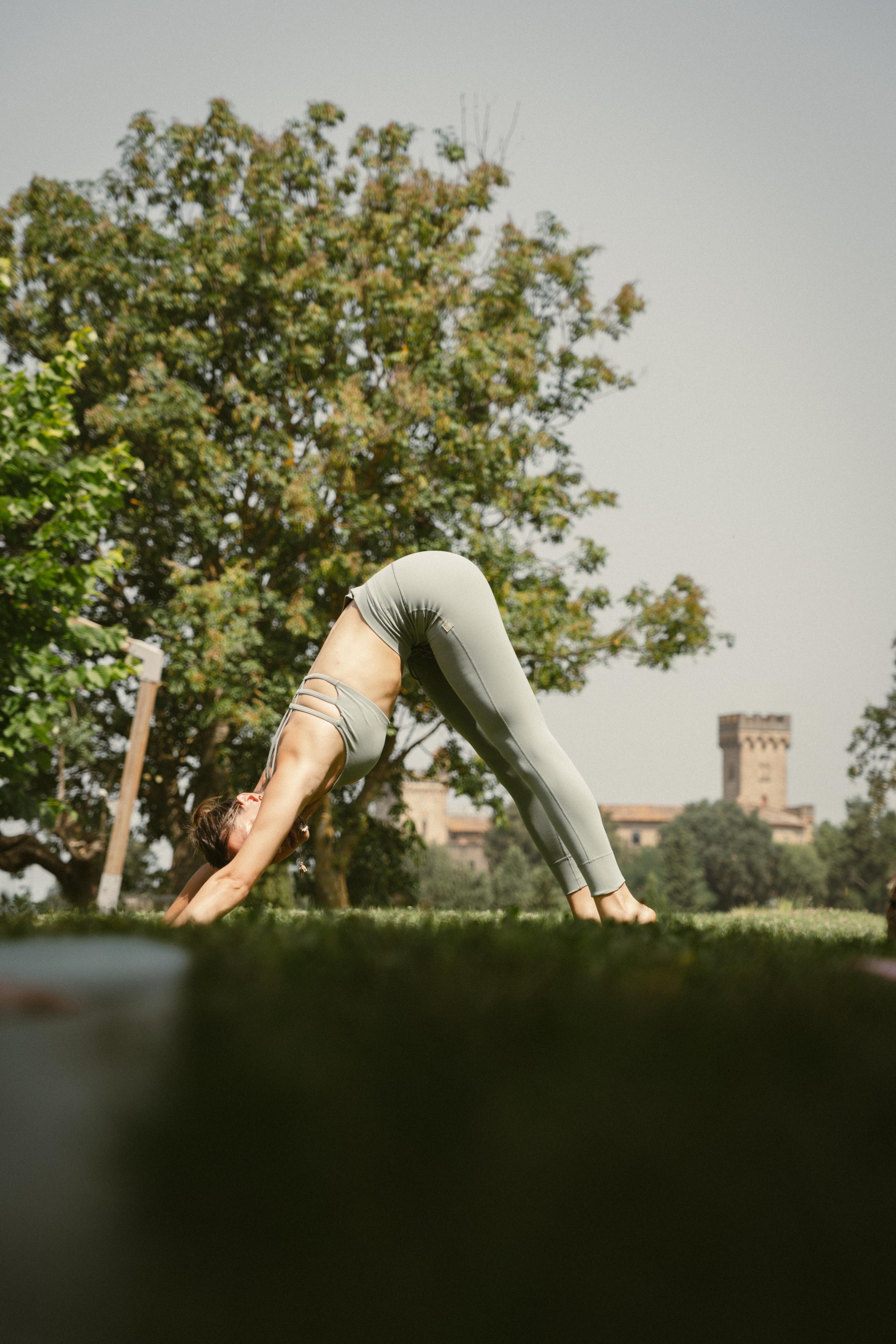 Una persona che fa yoga all'aperto in posizione del cane rivolto verso il basso, con uno sfondo di alberi e un edificio antico.