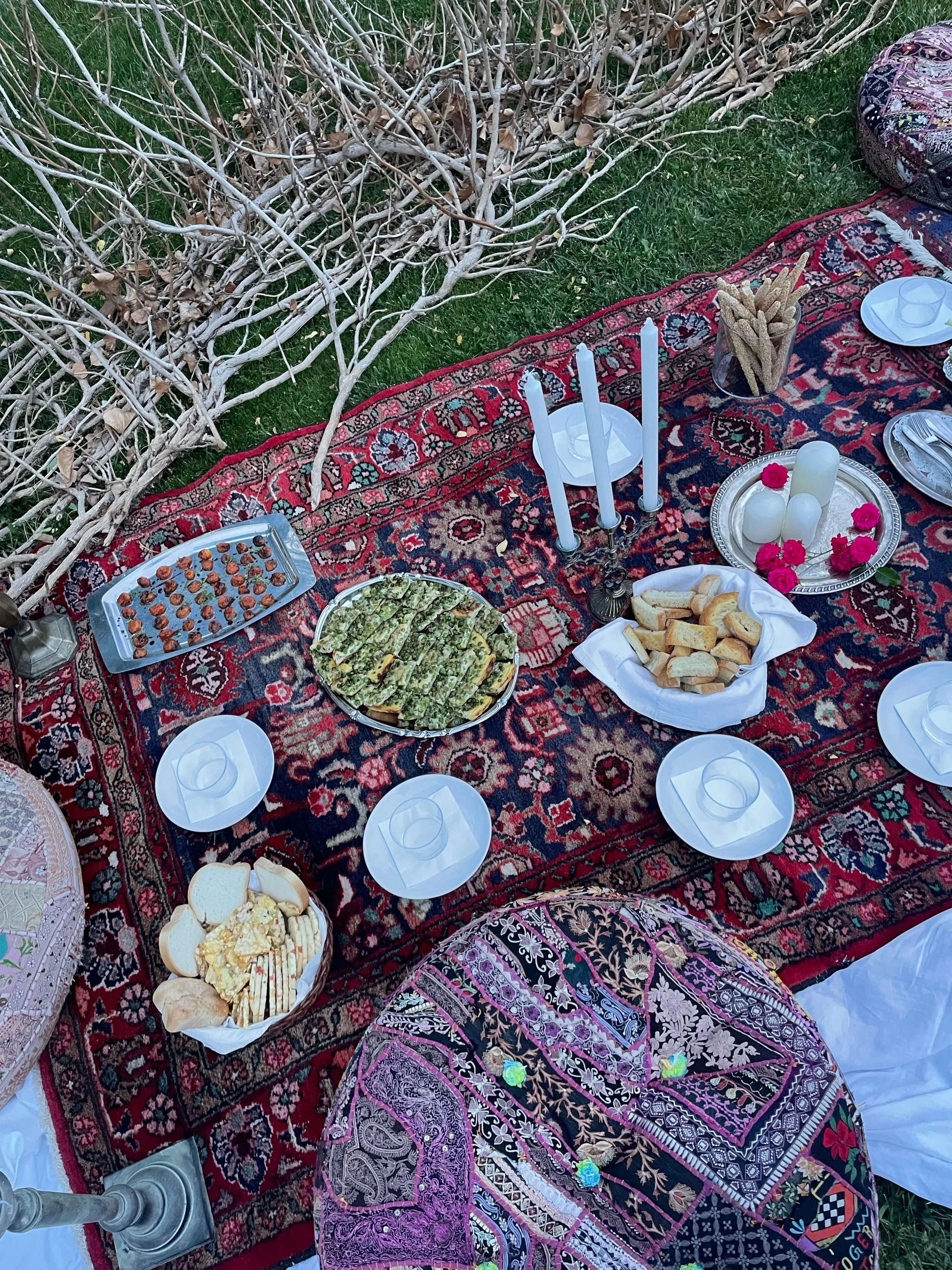 Un picnic appare su un tappeto di tessuto orientale decorato, con piatti di cibo come pane, formaggi, frutta e dolci, tra candele e bicchieri, ambientato all'aperto con erba verde e rami secchi.