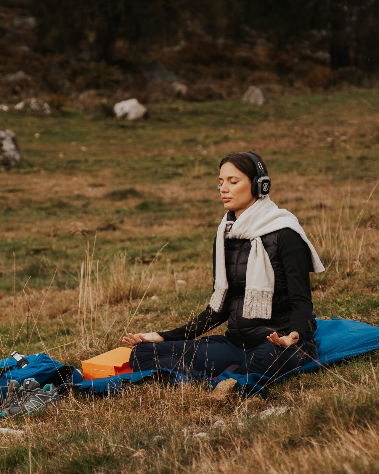 Donna seduta in meditazione sulla natura, indossa cuffie e abbigliamento da escursione, circondata da erba e rocce.