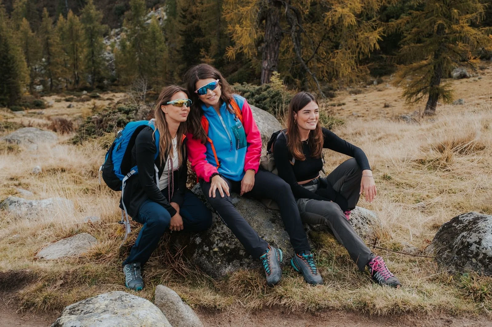 Tre donne sedute su un masso in un paesaggio montano con alberi e erba secca, indossano abbigliamento da escursionismo con zaini e occhiali da sole.