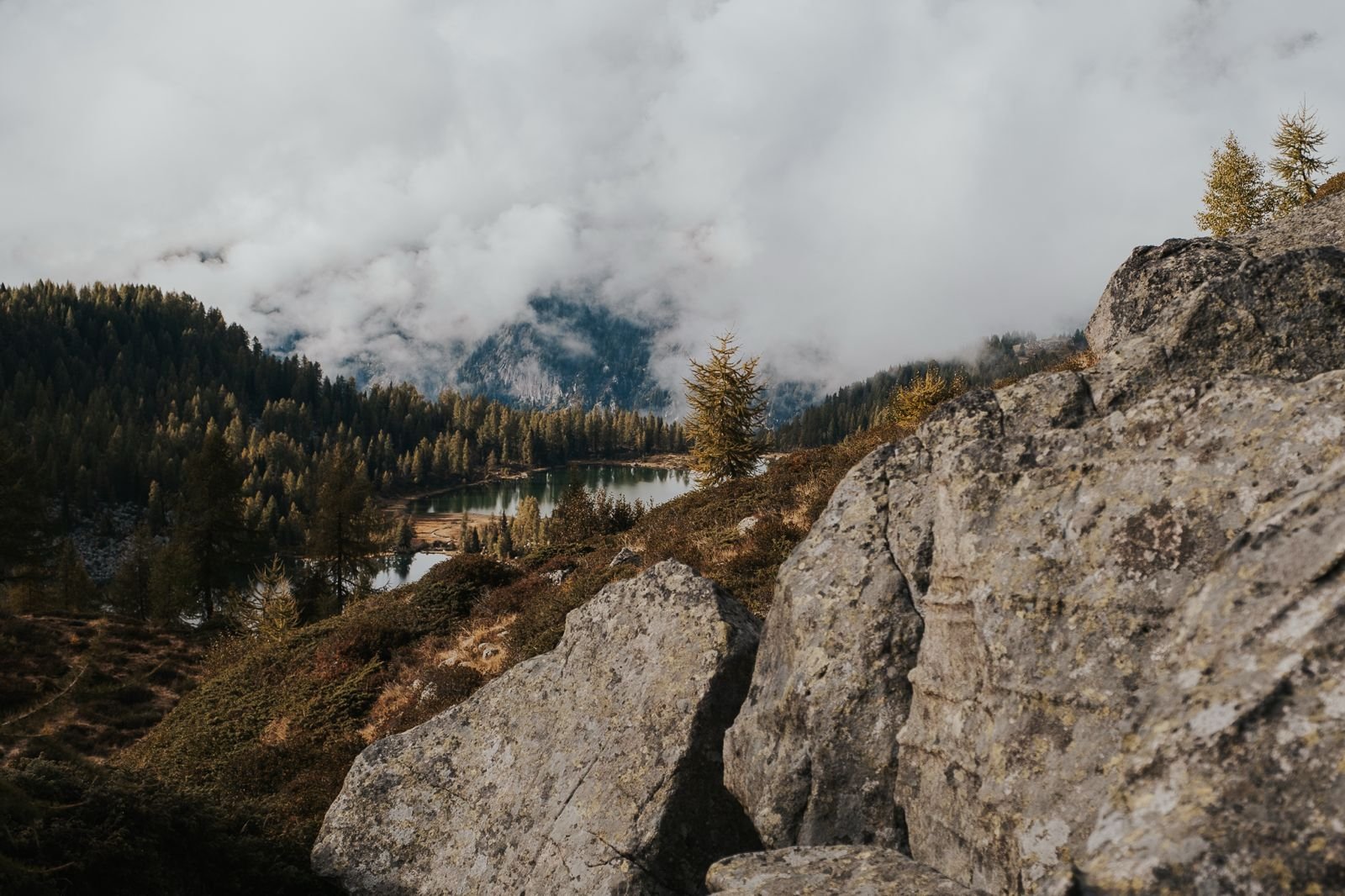 Paesaggio di montagna con laghetto, alberi e rocce in primo piano, nuvole e montagne sullo sfondo.