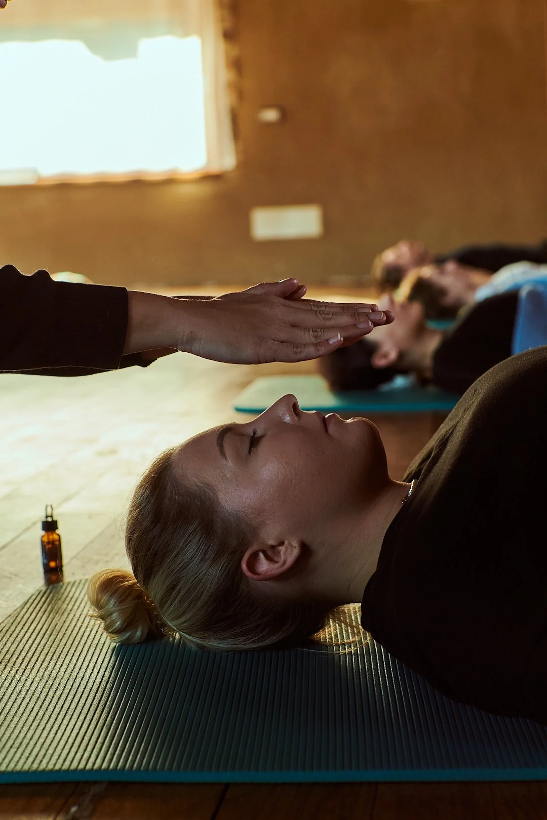 Donne che assistono a una lezione di yoga o meditazione, con una donna in primo piano che si trova sdraiata su un tappetino, in un ambiente interno con una finestra illuminata sullo sfondo.
