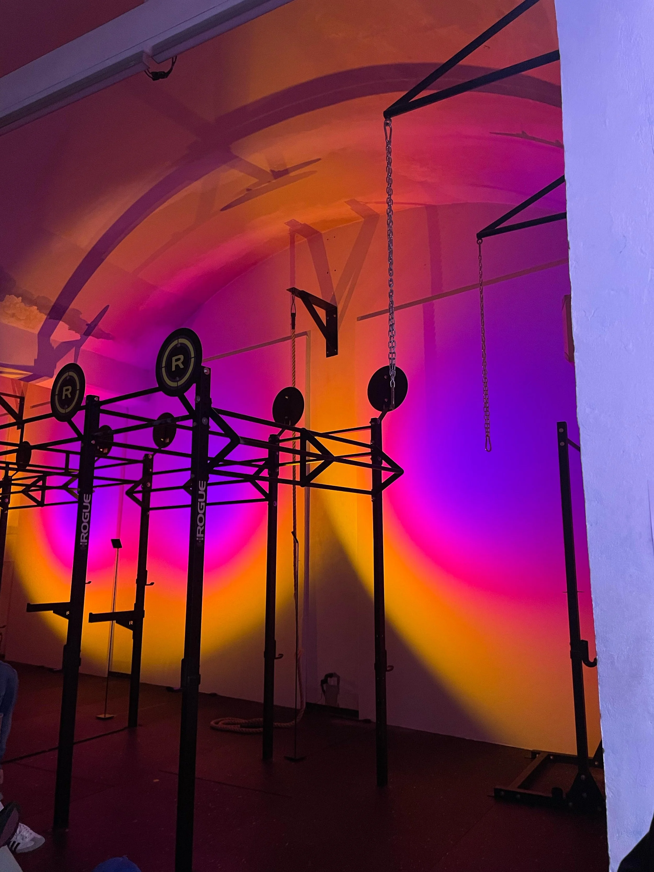 Struttura per allenamento CrossFit con barre, catene e attrezzature in un ambiente con pareti illuminate con luci di colore viola, rosa e giallo.