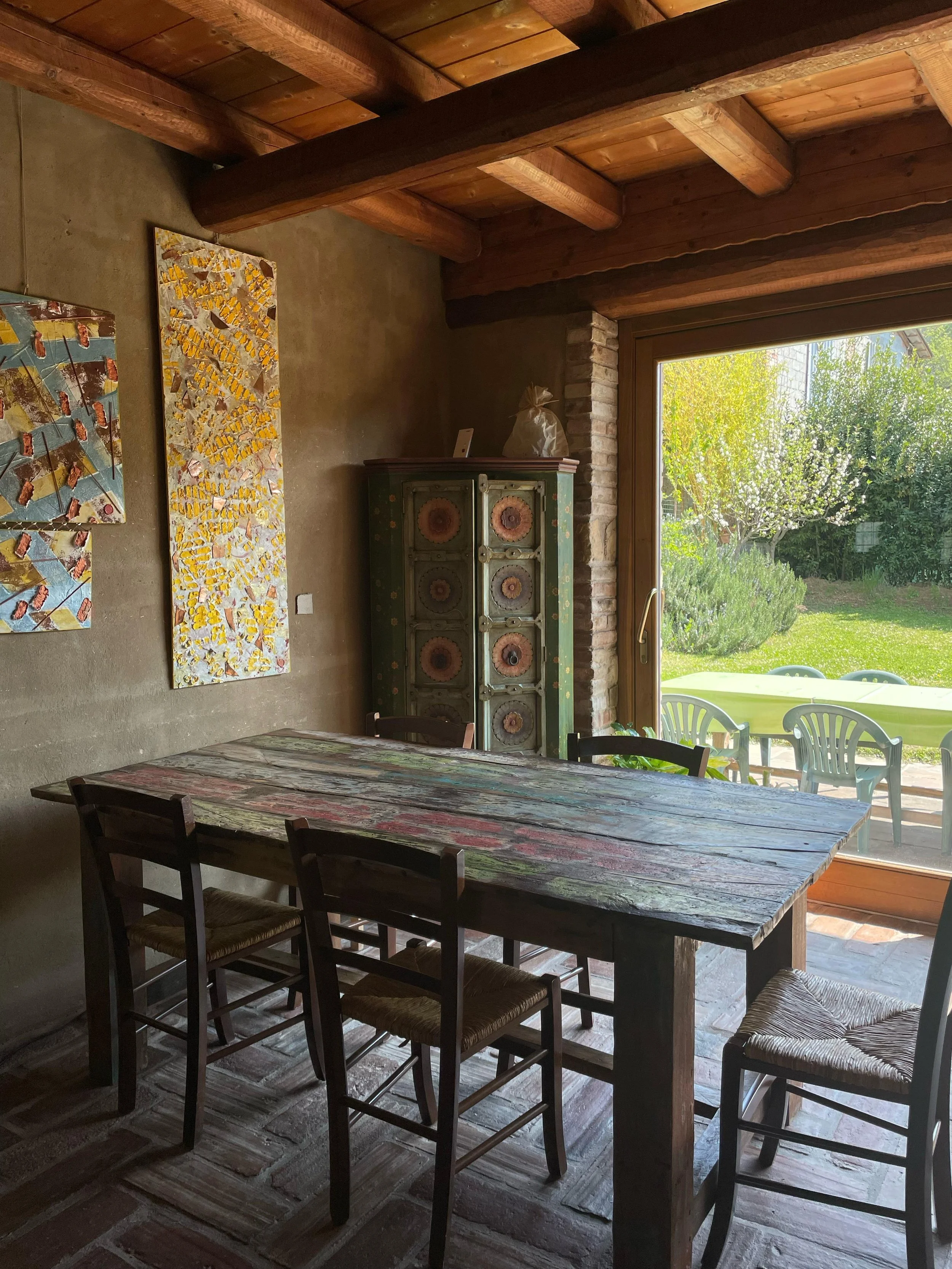 Sala da pranzo con tavolo di legno colorato, sedie di legno con sedili di paglia, muratura in mattoni e un armadio verde con decorazioni floreali, grande finestra che dà su un giardino con alberi e erba