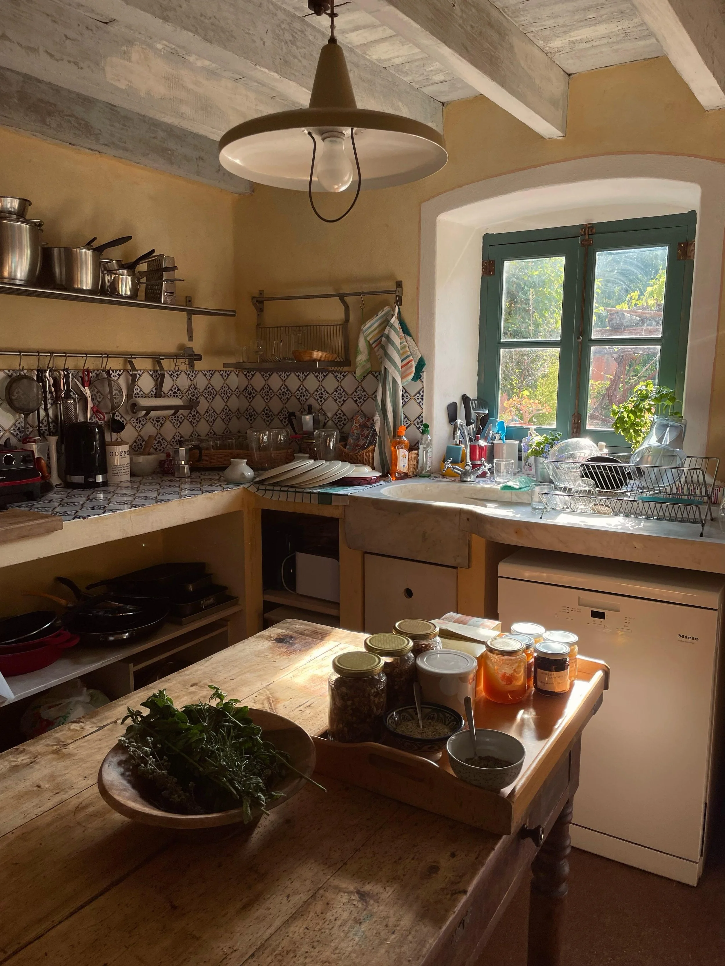 Una cucina rustica con finestra, piano di lavoro con vari utensili, barattoli e piante, lampadario in stile vintage.