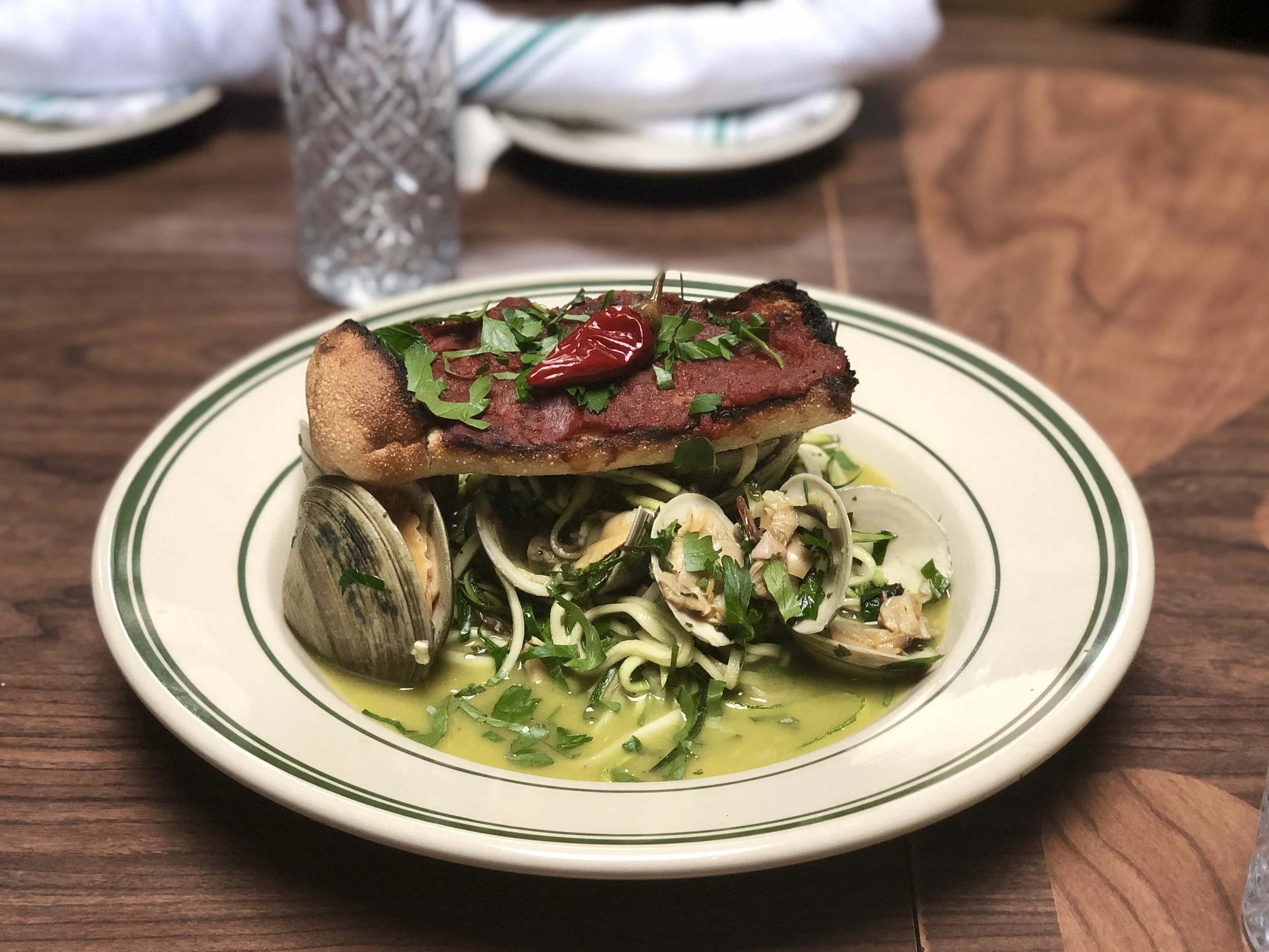 Seafood pasta with clams, topped with grilled meat, herbs, and red chili on a white plate with green stripes.
