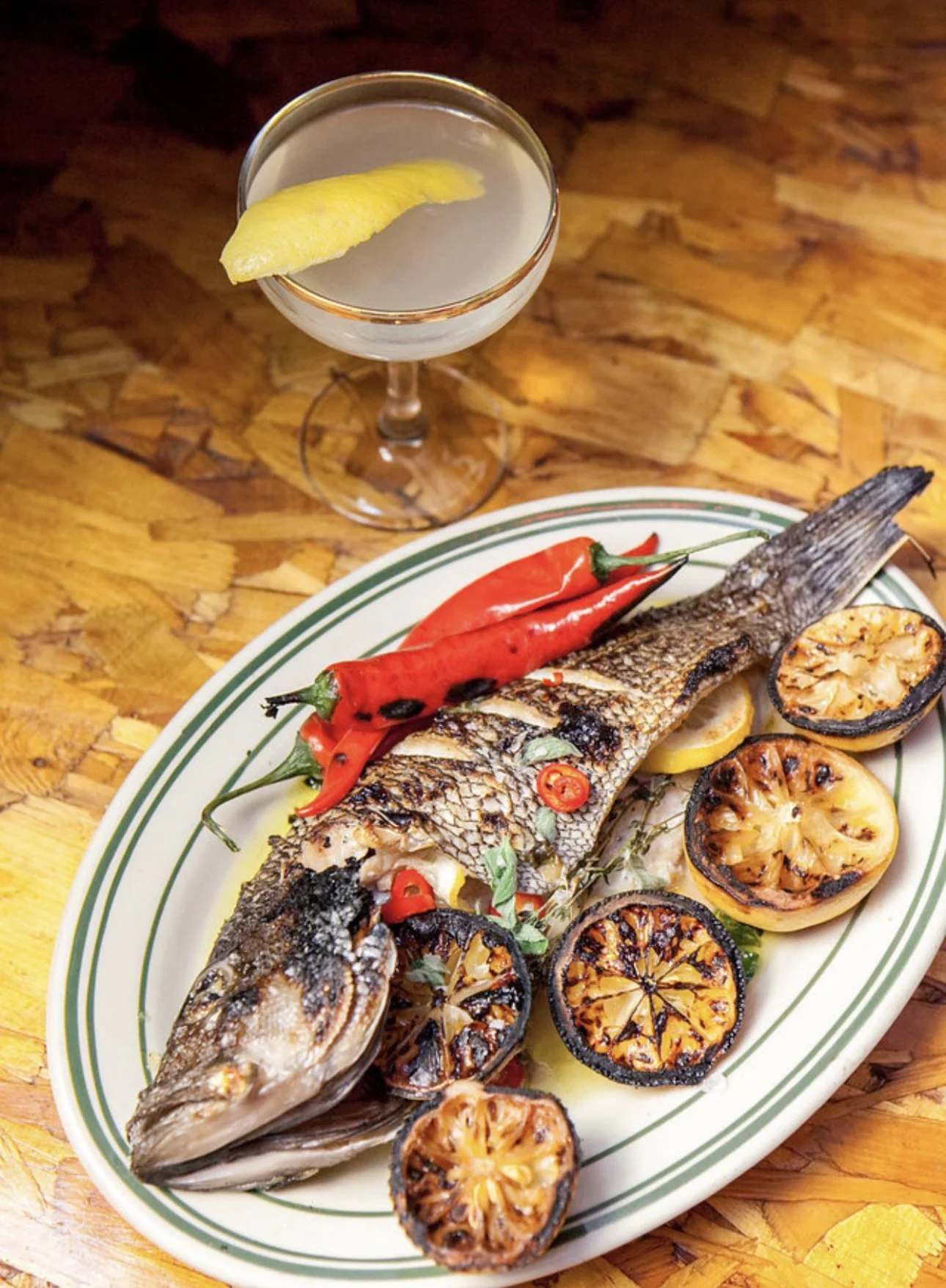 A plate with grilled whole fish garnished with grilled lemon slices and red chili peppers, accompanied by lettuce and herbs, on a wooden table. A cocktail with a lemon wedge is in the background.