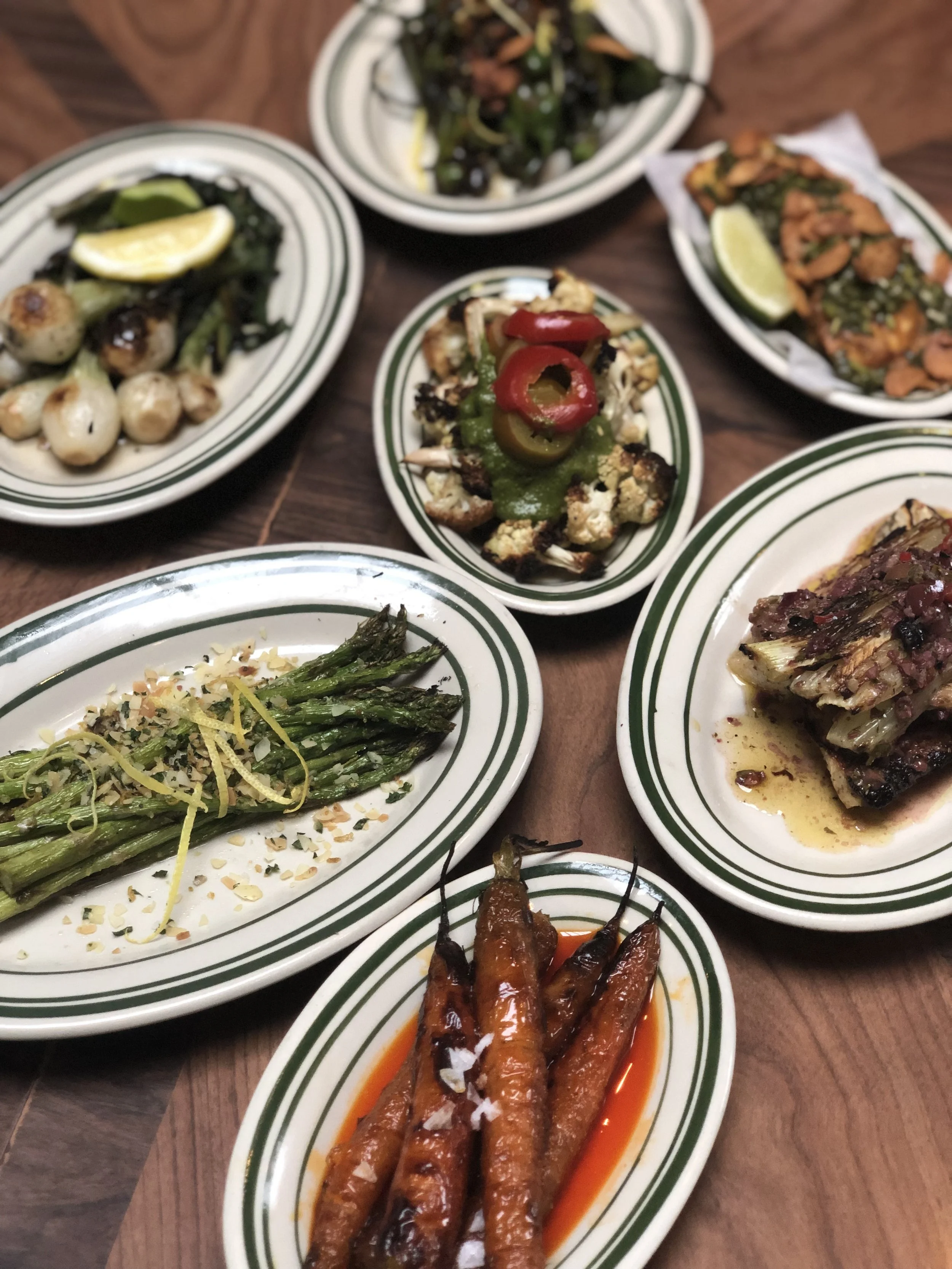 Assorted Middle Eastern dishes on green-and-white striped plates, including grilled vegetables, stuffed grape leaves, roasted carrots, tabbouleh, and dishes with lemon wedges.
