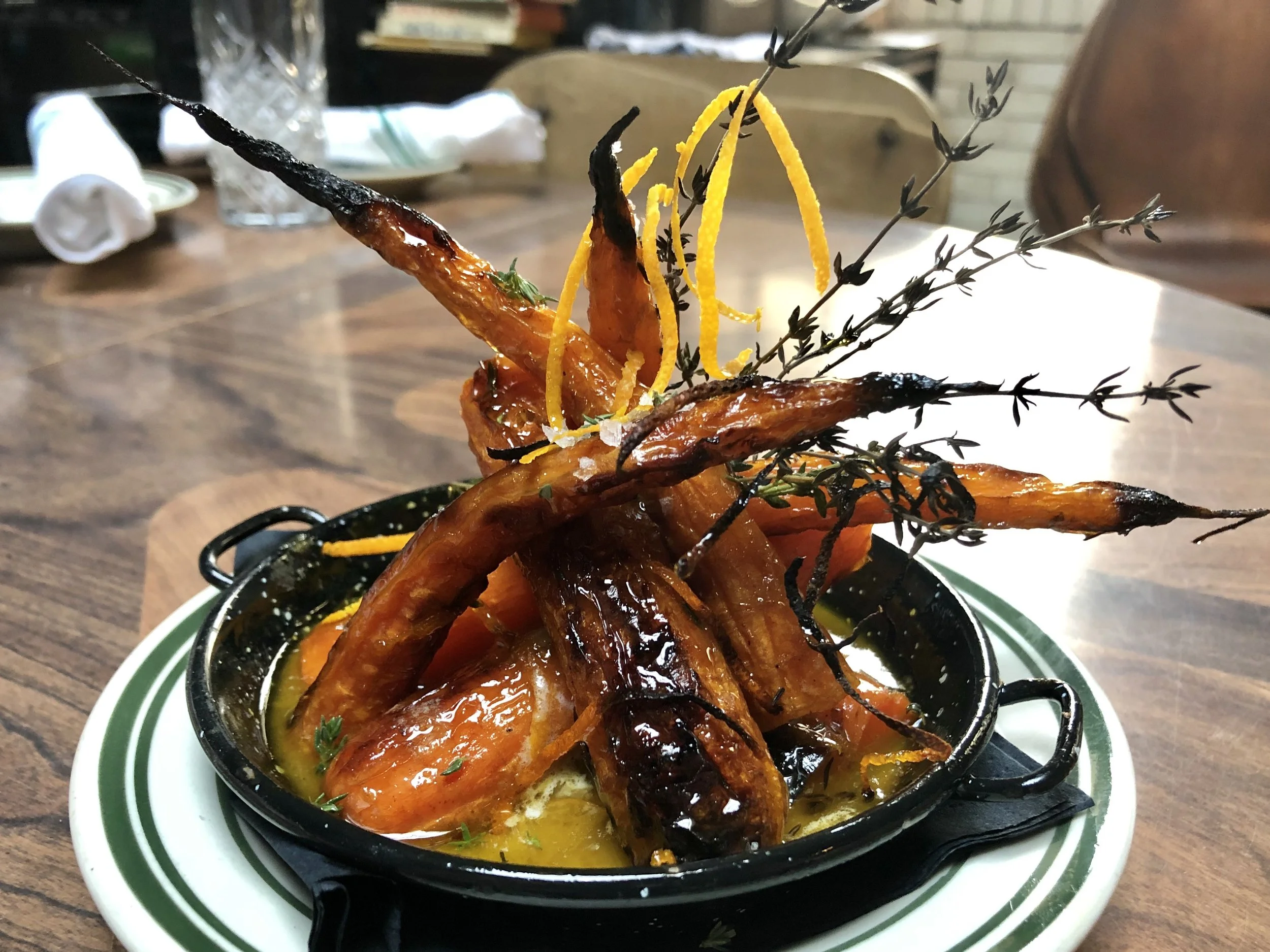 A dish of roasted carrots garnished with sprigs of thyme and lemon peel, served in a black skillet on a white plate with a green striped rim.