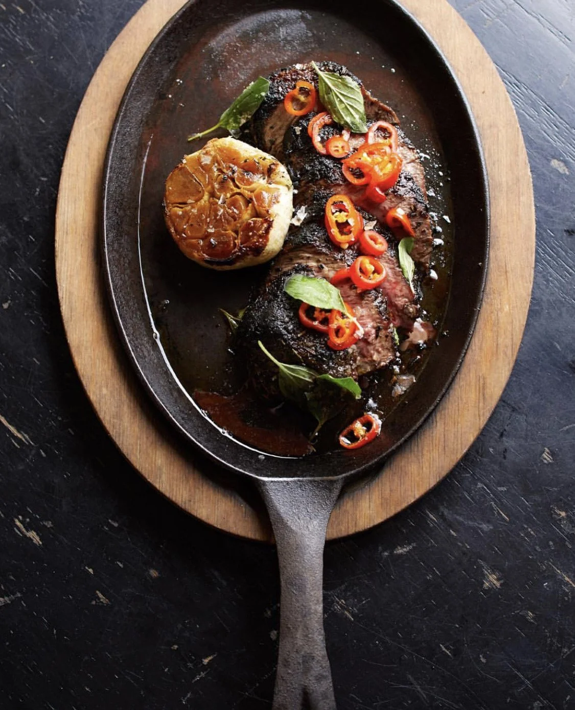 A cooked beef steak topped with sliced red chili peppers and fresh basil, served with roasted garlic and herbs on a black cast iron skillet placed on a wooden board.