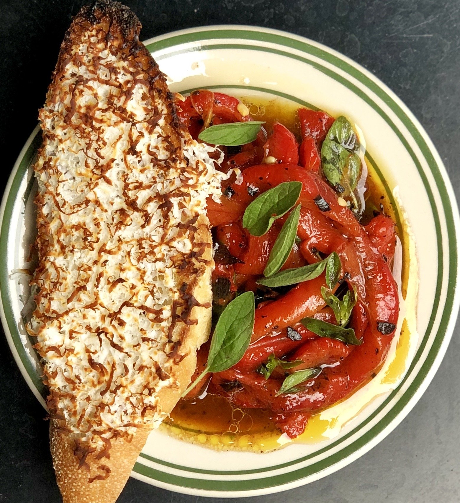 Slice of toasted bread topped with melted cheese and herbs served with roasted red peppers and fresh basil leaves.