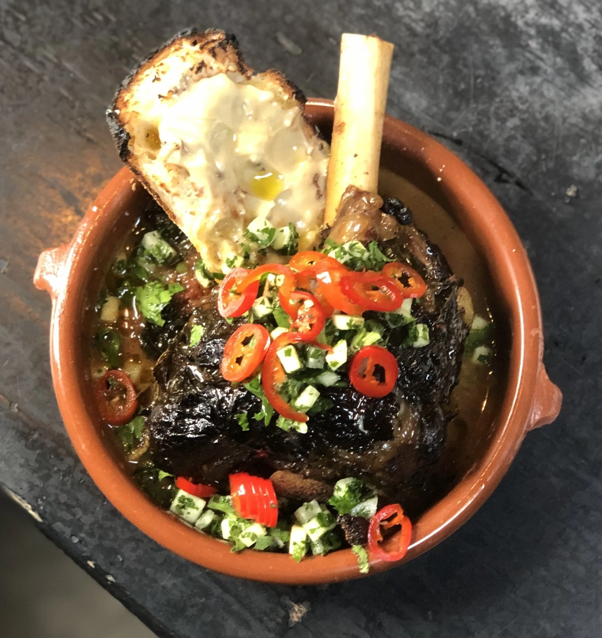 A bowl of spicy meat stew garnished with chopped green onions and sliced red chili peppers, with a piece of toasted bread and a marrow bone on top.