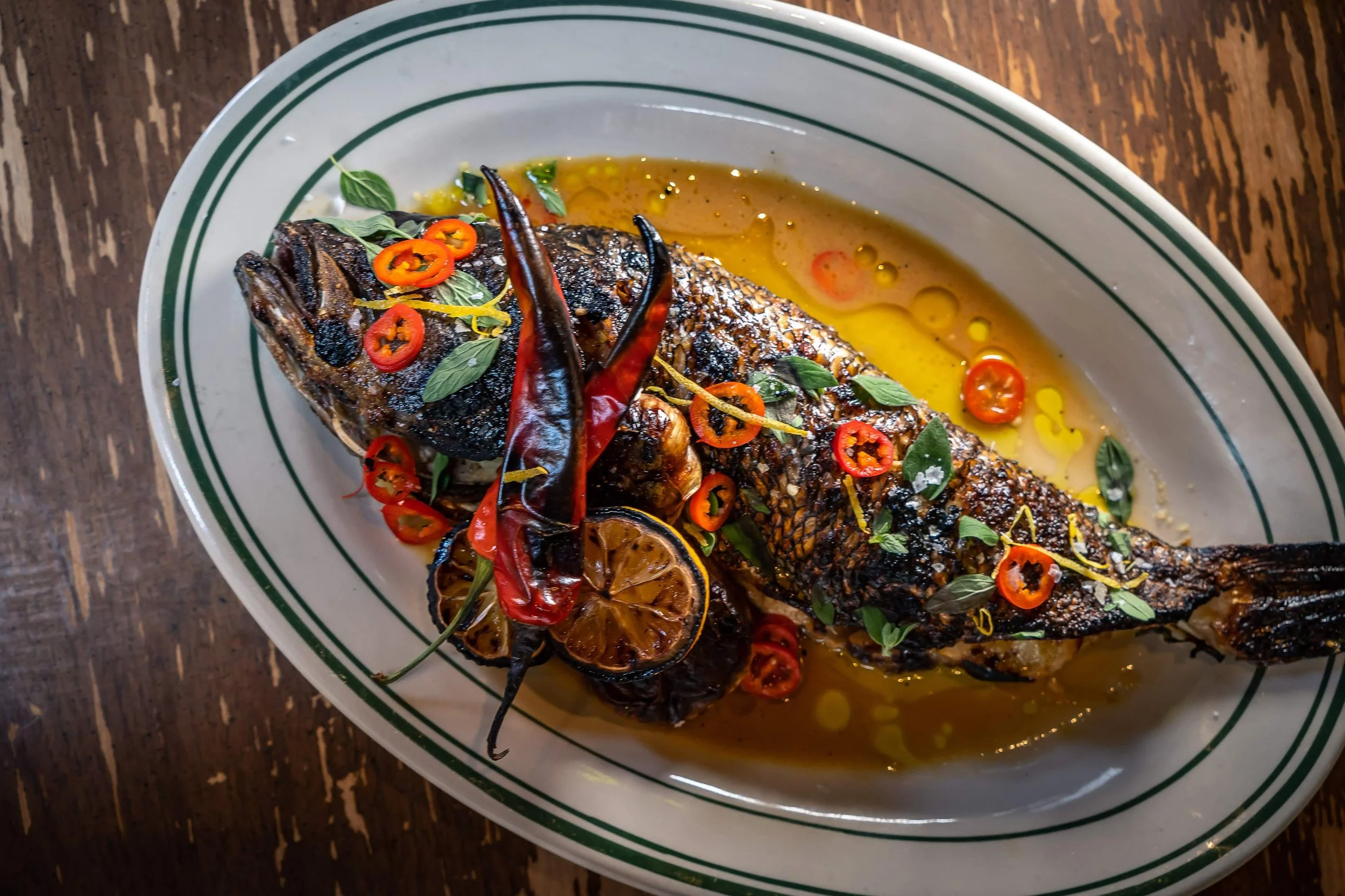 Grilled whole fish garnished with red chili peppers, lemon slices, and fresh herbs, served on an oval plate with sauce.