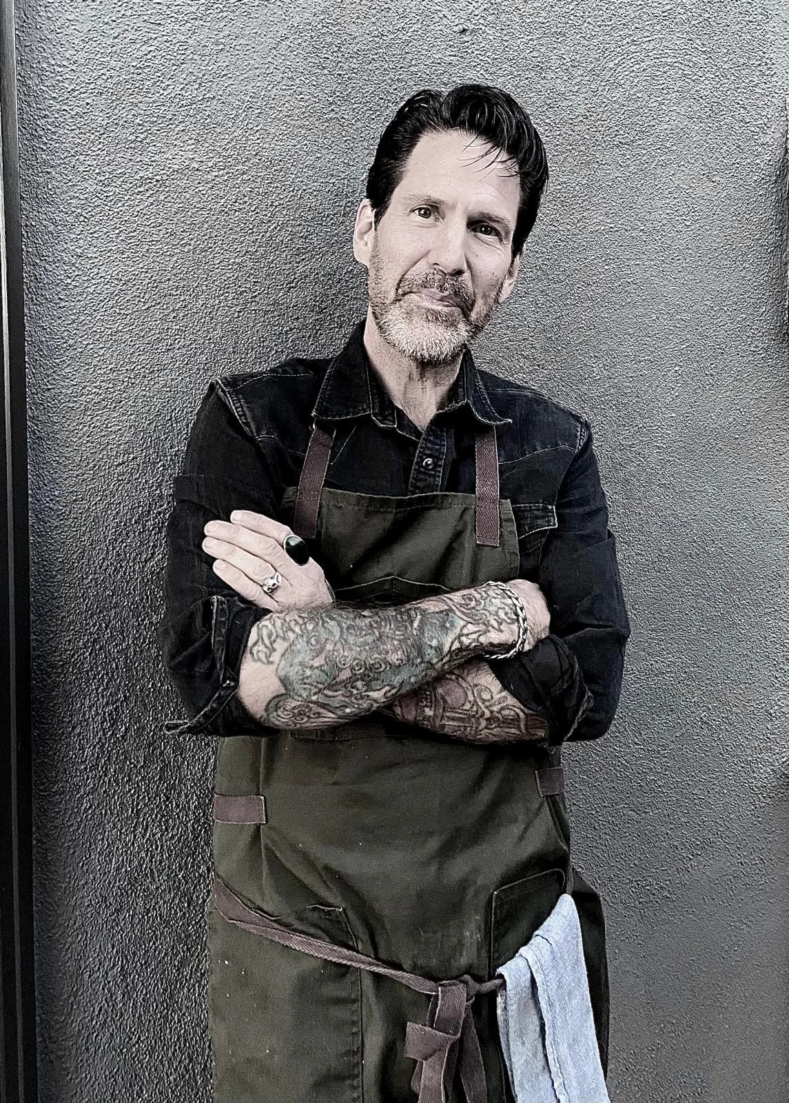 A man with dark hair, a beard, and tattoos on his arms standing with arms crossed wearing a black shirt, an apron, and a ring on his finger against a textured wall.