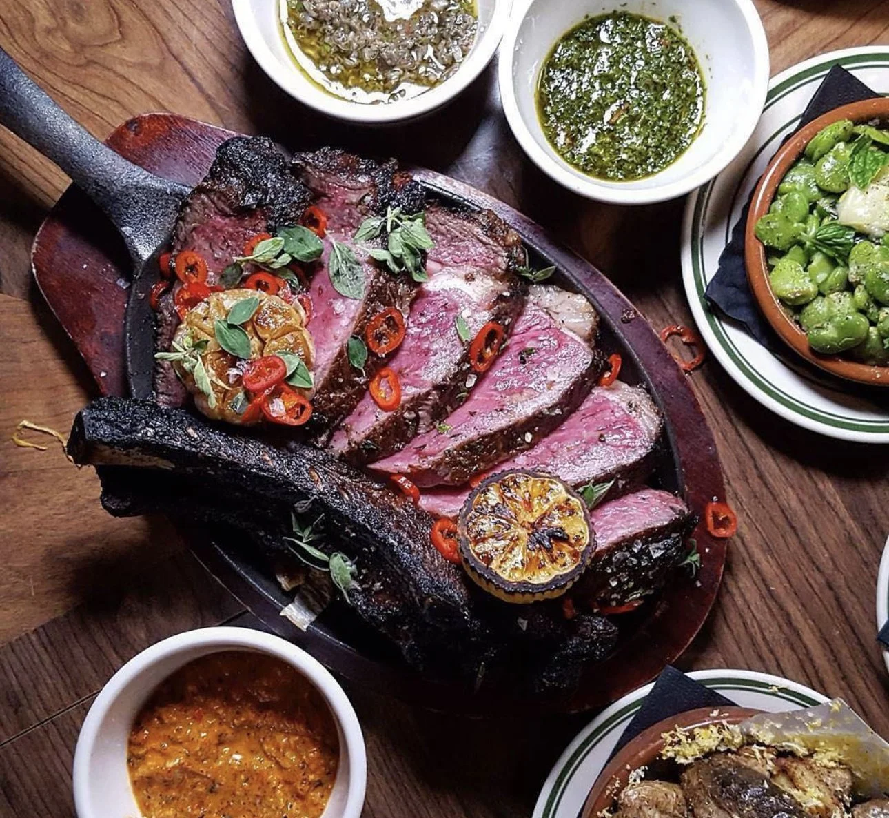 Slices of cooked beef steak topped with red chili slices, herbs, and grilled lemon on a wooden table with various sauces and side dishes.