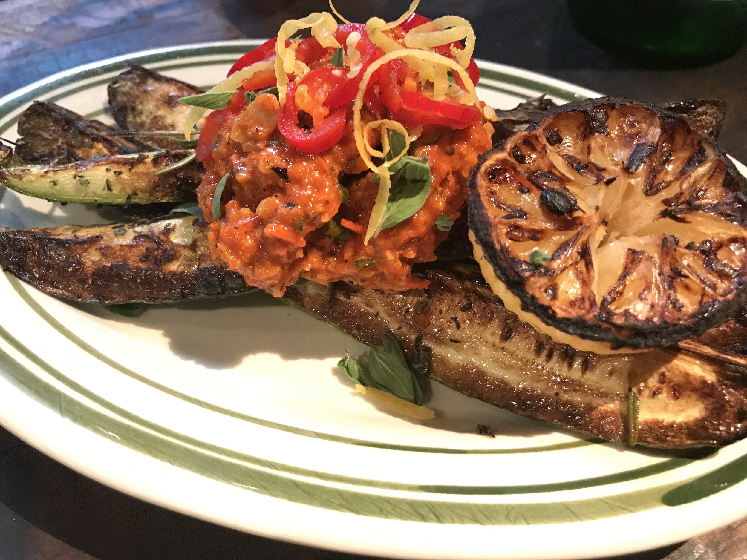 Grilled eggplant topped with lemon slices, served with roasted vegetables and tomato salsa on a white plate with green stripes.