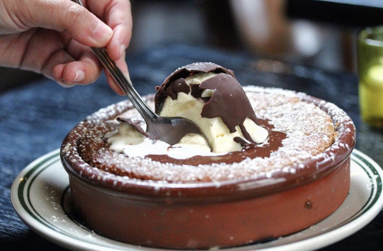 Dessert with ice cream, chocolate and vanilla, served in a brown dish with powdered sugar, with a hand using a spoon to scoop some.