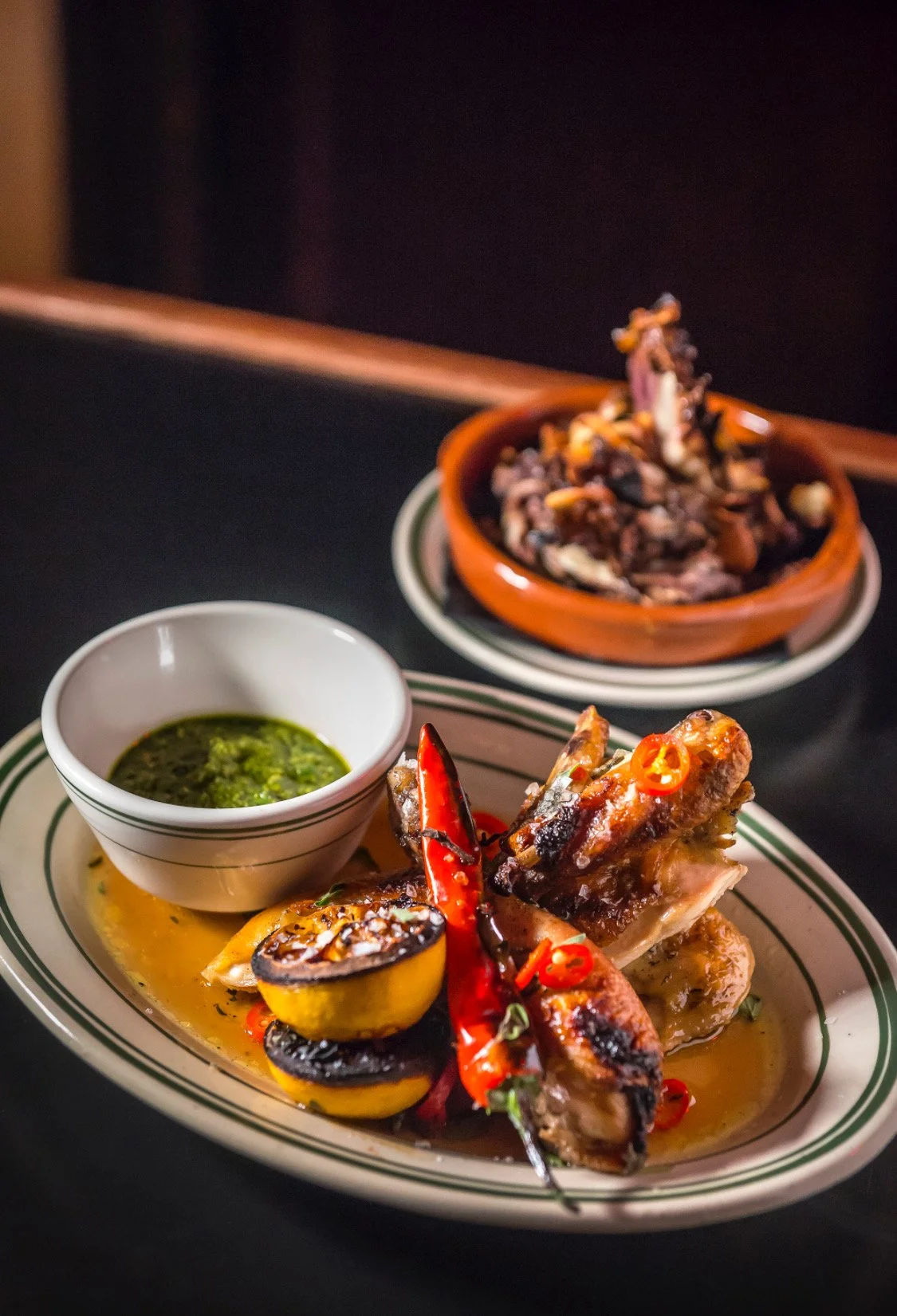 A plate of grilled chicken with red chili peppers, grilled lemon, and green sauce, accompanied by a bowl of salsa or sauce, with another plate of cooked meat in the background.