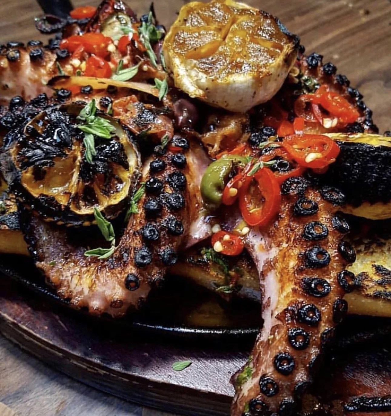 Grilled octopus tentacle garnished with red chili slices, lemon, and herbs, served with roasted vegetables.
