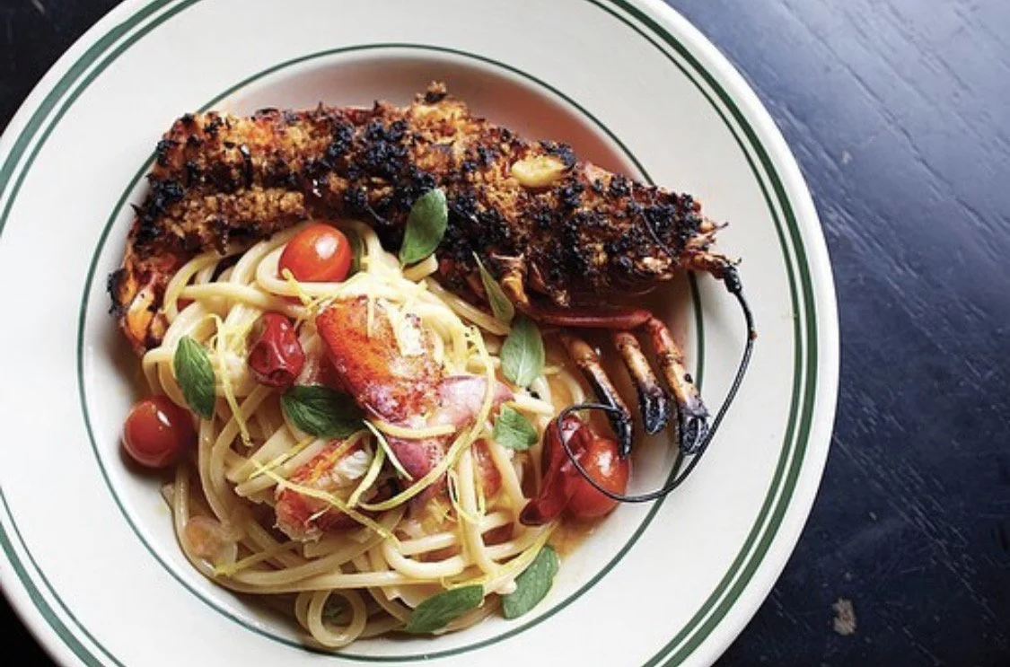 Grilled lobster with a side of pasta with cherry tomatoes and fresh herbs on a white plate with green stripes.