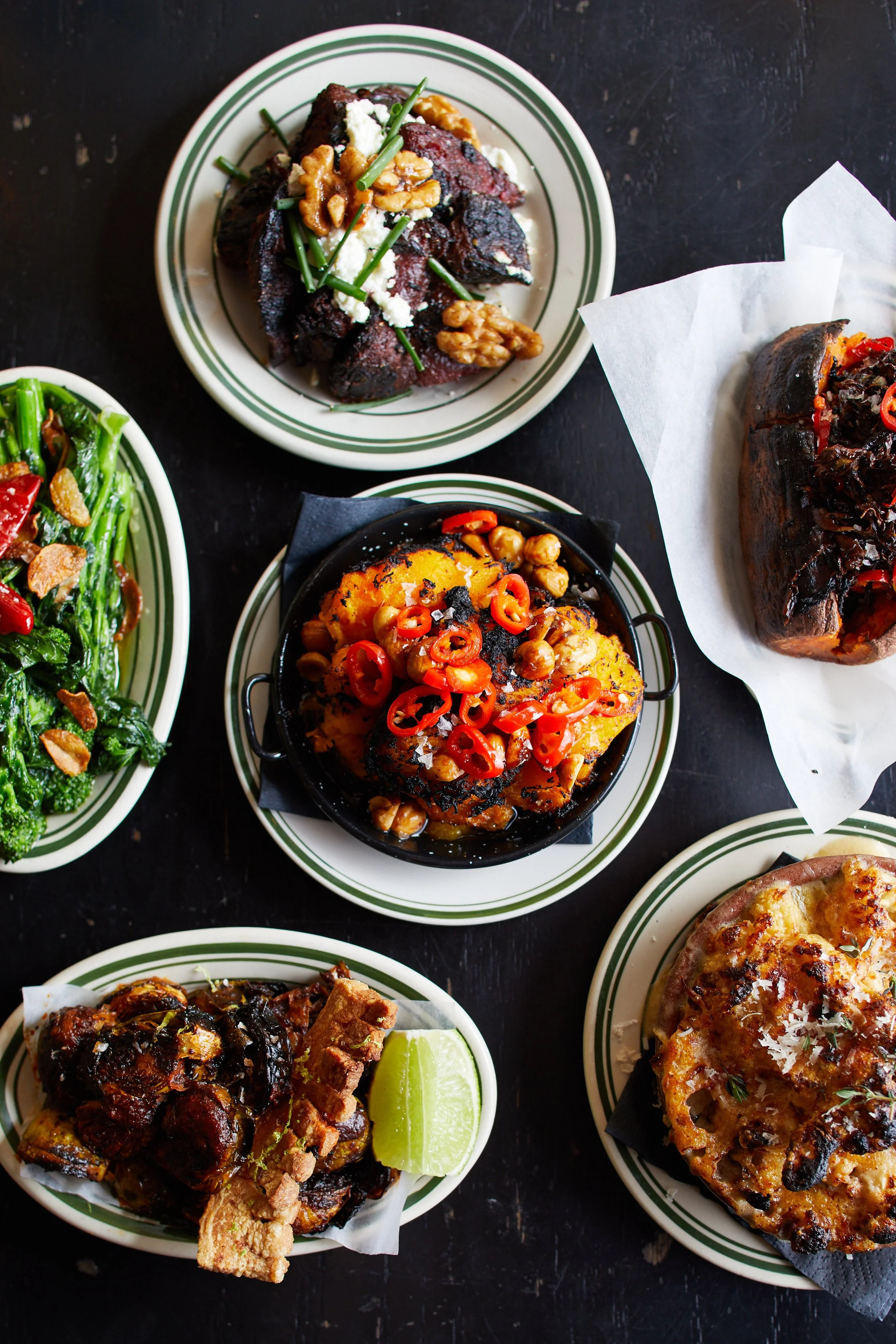 Variety of Mexican dishes on striped plates, including grilled meat, roasted vegetables, volcano dish topped with chili peppers, and pizza with cheese and herbs, on a dark surface.
