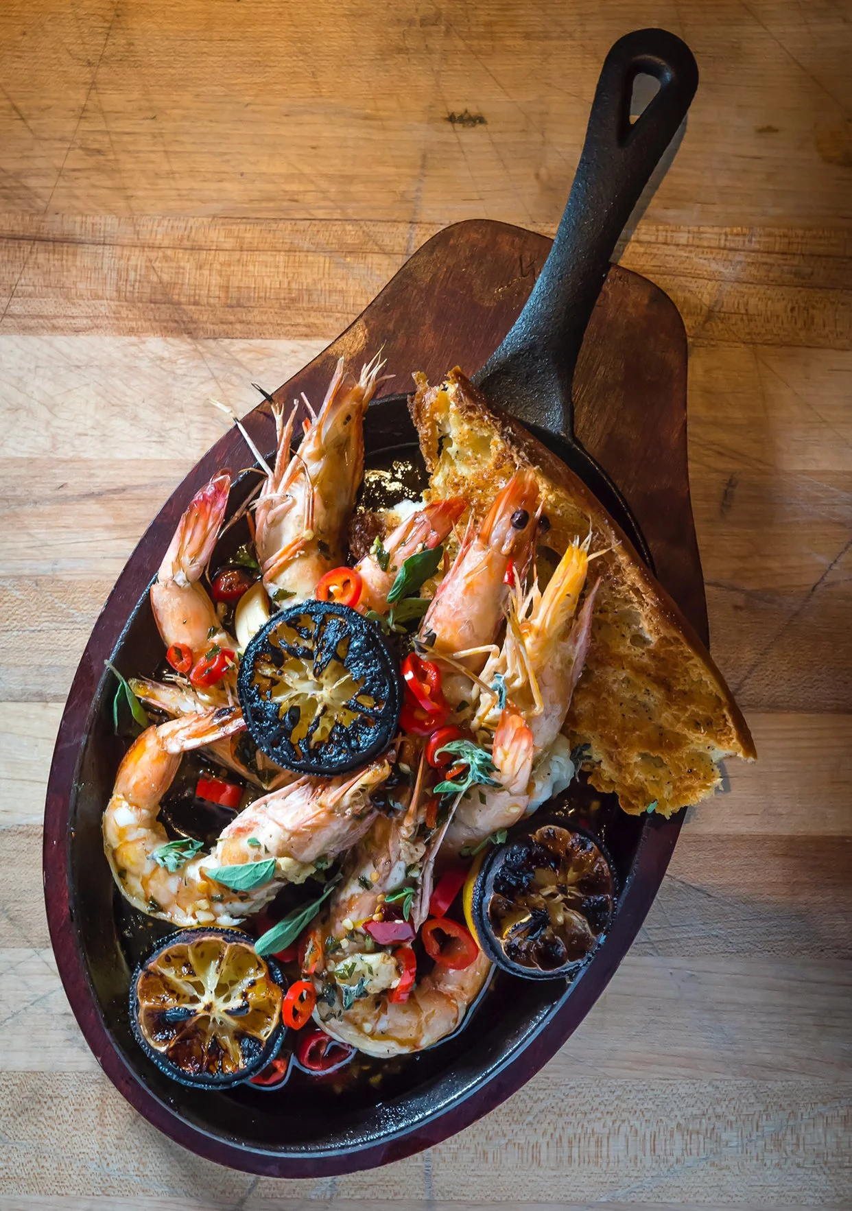 Fresh shrimp with grilled lemon slices, red chili peppers, and a piece of toasted bread in a black skillet