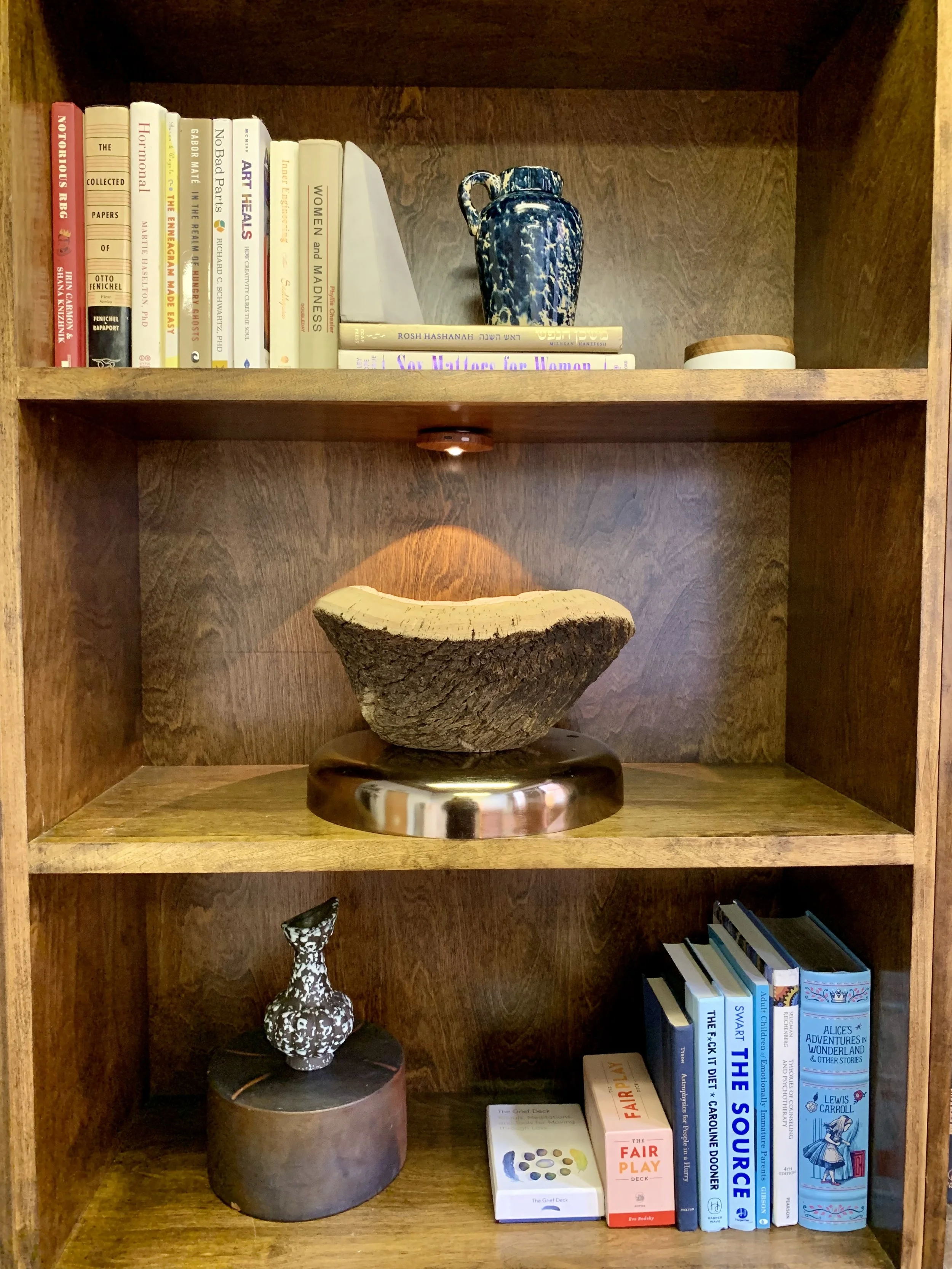 A wooden bookshelf with three shelves containing books, decorative objects, and a piece of wood art. The top shelf has a blue ceramic vase, some books, and a small round container. The middle shelf has a piece of wood art on a stand. The bottom shelf has a small decorative vase, a box of playing cards, and several books including "The Source" and "The Fairy Deck".