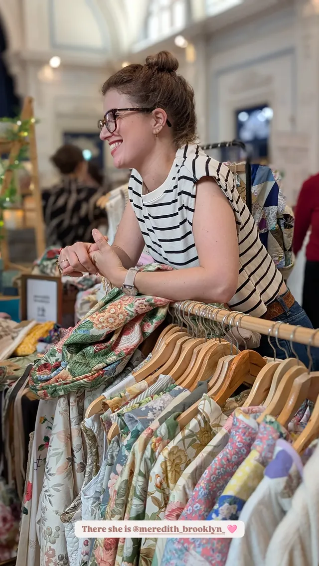 designer leans on rail of handmade clothing smiling at customers