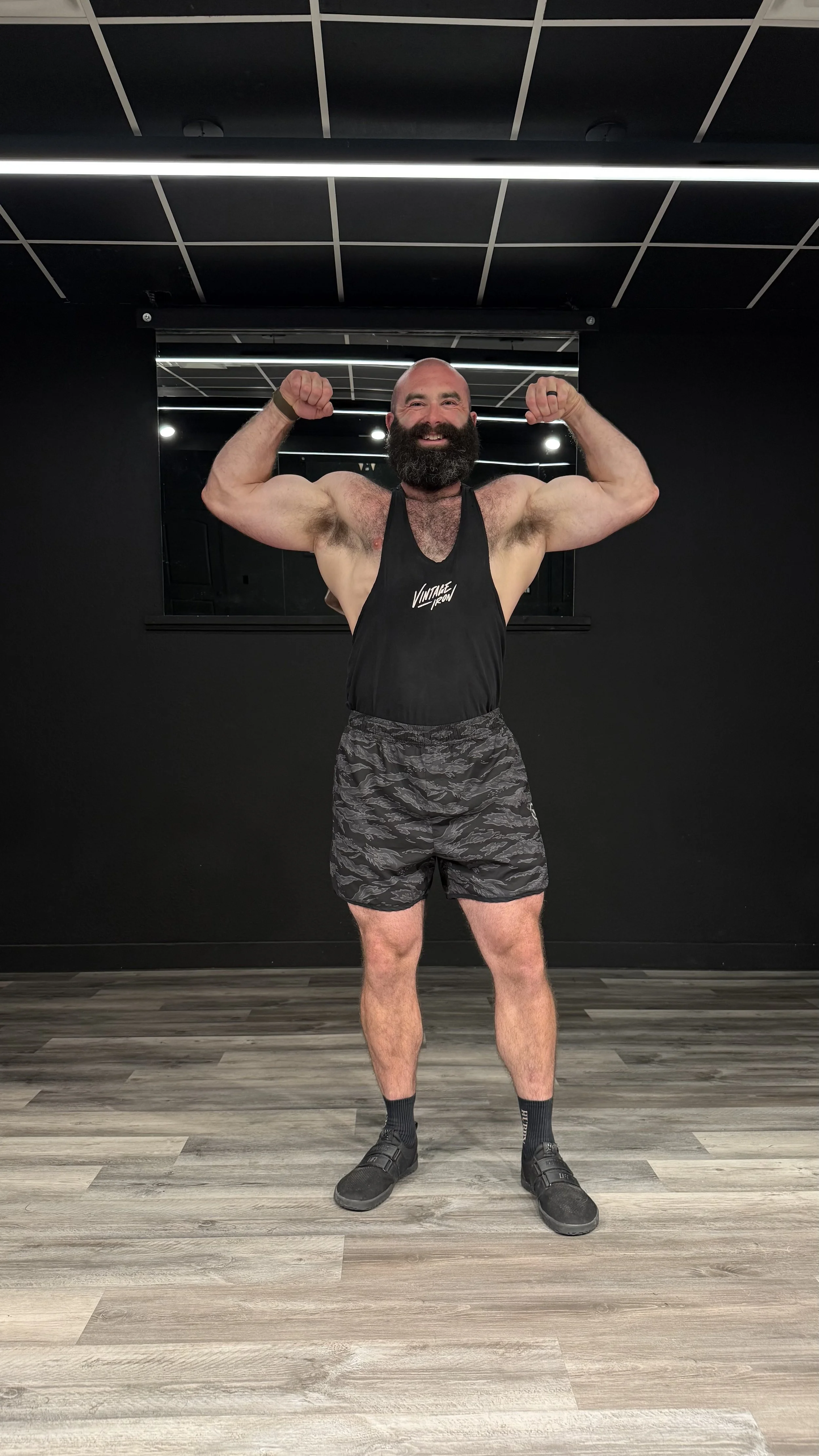 A man with a bald head and a full beard, smiling and flexing his biceps, standing in a gym with dark walls, a black mirror behind him, a wooden floor, and wearing a black tank top, camouflage shorts, black socks, and black shoes.