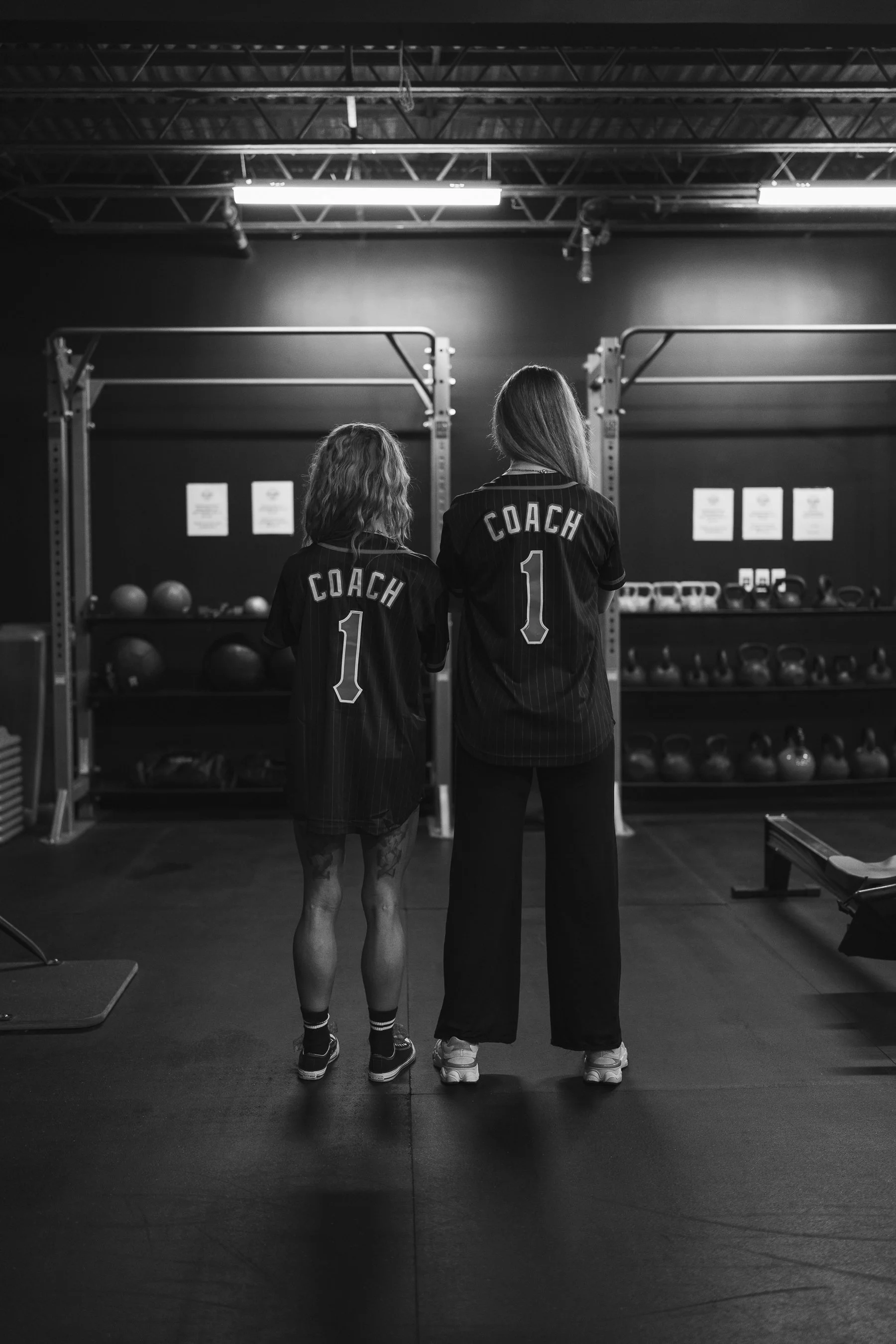 Two women in a gym, both wearing sports jerseys labeled 'COACH' with the number 1, standing side by side and facing away from the camera. The gym has exercise equipment like medicine balls and kettlebells in the background, with an industrial ceiling and bright lighting.