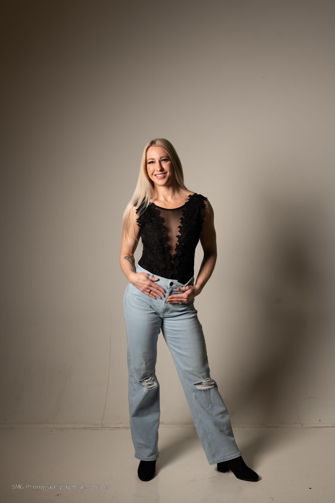 A woman with long blonde hair wearing a black lace top and light blue ripped jeans, standing against a plain beige background, smiling at the camera.