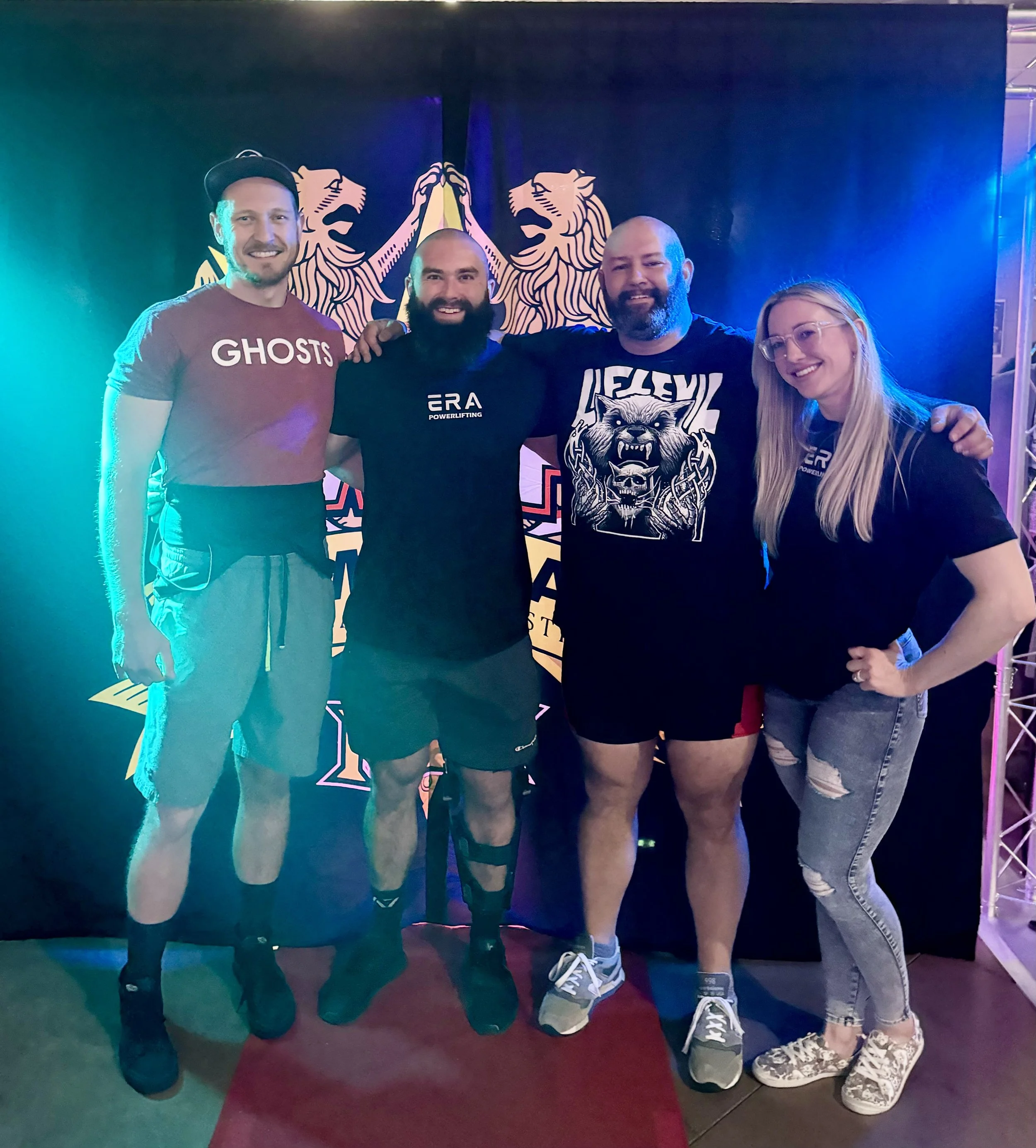 Four people standing together in front of a backdrop with two illustrated lions. The group includes two men and one woman. They are smiling and posing for the photo, dressed casually. The setting appears to be a social gathering or event with colorful lighting.