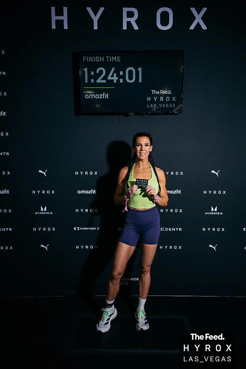 A woman standing on a treadmill at the Hyrox fitness event in Las Vegas, holding a medal, smiling, with a digital clock displaying 1:24:01 behind her.