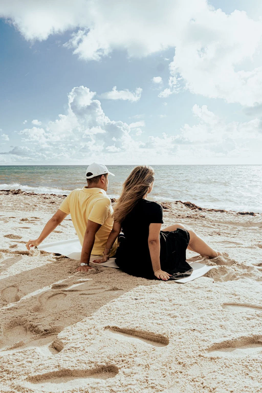 A man and woman sitting on the sandy beach looking out at the ocean under a partly cloudy sky.