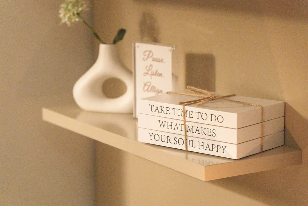 A white shelf holding a white vase with a flower, a framed quote, and three stacked hardcover books tied with string. The books have a quote on the side that reads, "Take time to do what makes your soul happy."
