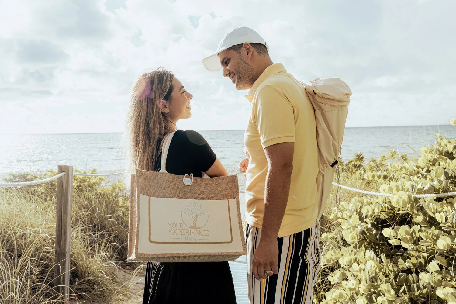 A woman and a man standing close on a beach, smiling at each other. The woman has a tote bag with the words 'Your Experience' and a tree logo. The man is wearing a yellow polo shirt, striped pants, and has a backpack, with the ocean and sky in the background.
