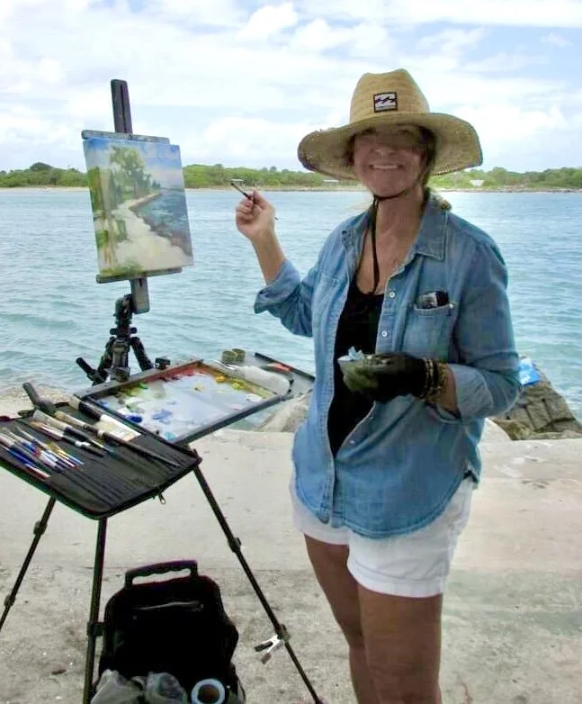 Image of Michaelann Bellerjeau, standing on a beach with a painting palette, easel, and brushes, holding a canvas and a palette while smiling at the camera, wearing a large straw hat, denim shirt, and white shorts with the ocean in the background.