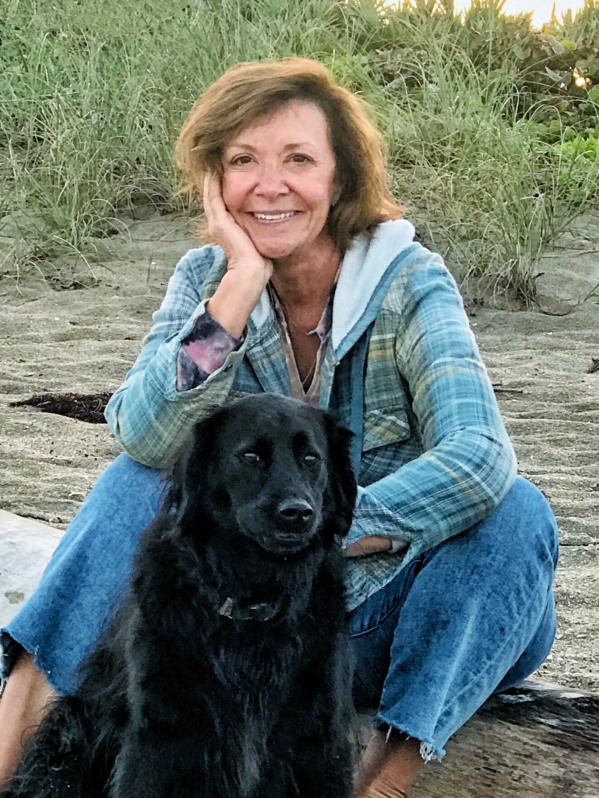 Image of Michaelann Bellerjeau, a woman with brown hair sitting on a wooden log on the beach, smiling, with a black dog sitting near her. The woman is wearing a blue plaid jacket and jeans, and the background features grassy dunes and a sunset.