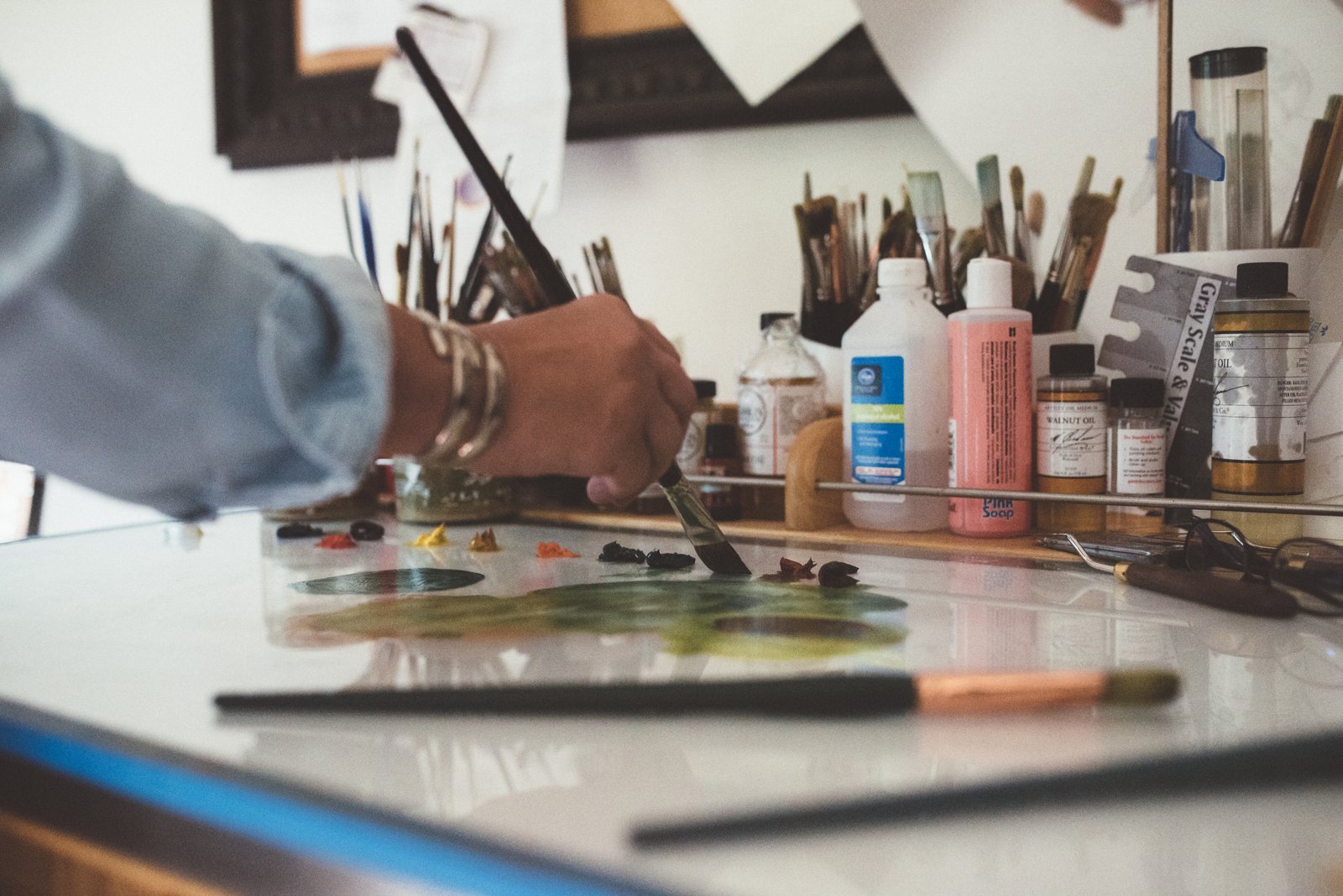 Artist mixing paints on a palette at an art station with various bottles, brushes, and tools in the background.