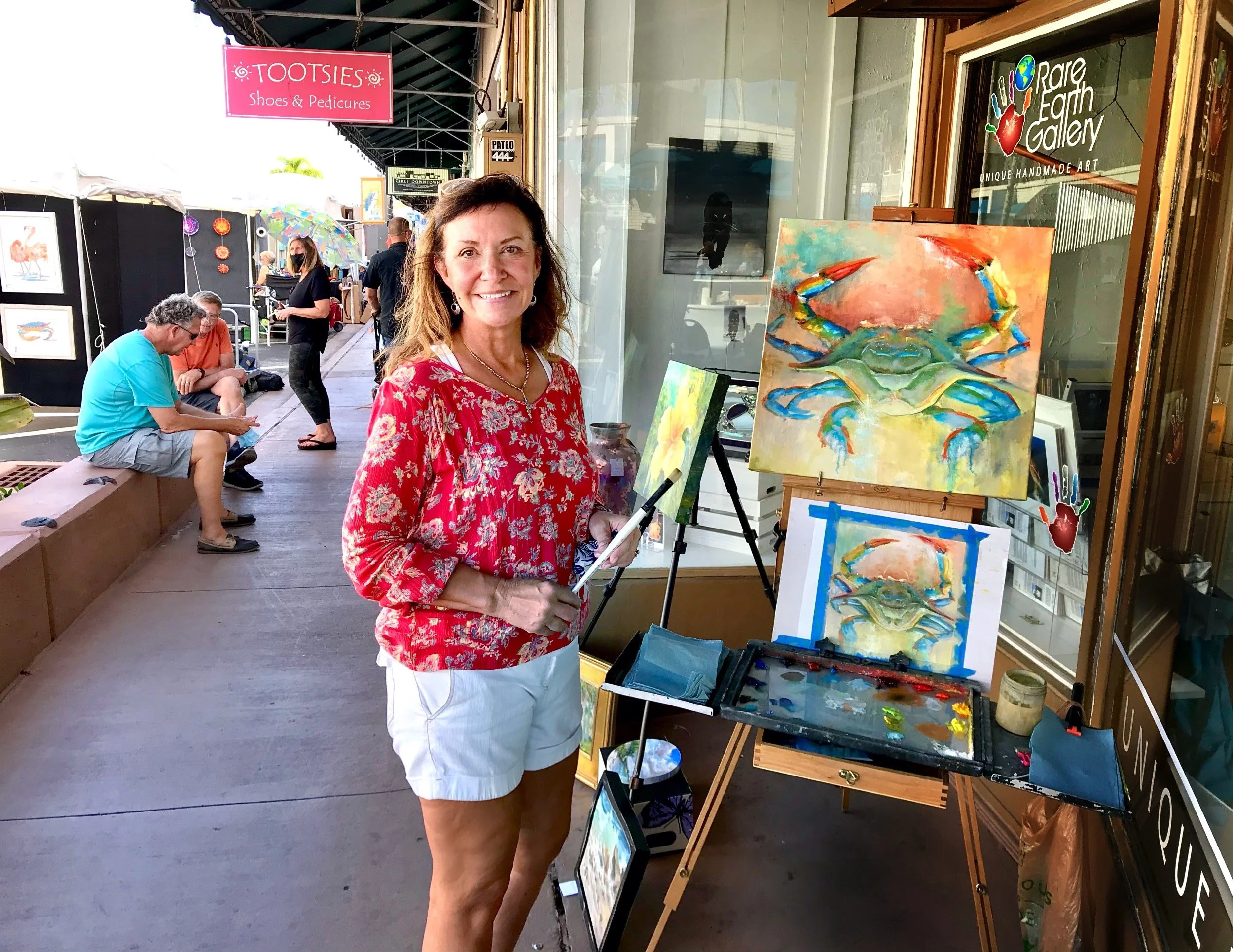 Image of Michaelann Bellerjeau, a woman painting a colorful crab on an easel outside an art gallery, Rare Earth Gallery in Stuart, Florida, with festivalgoers sitting and walking on the street.