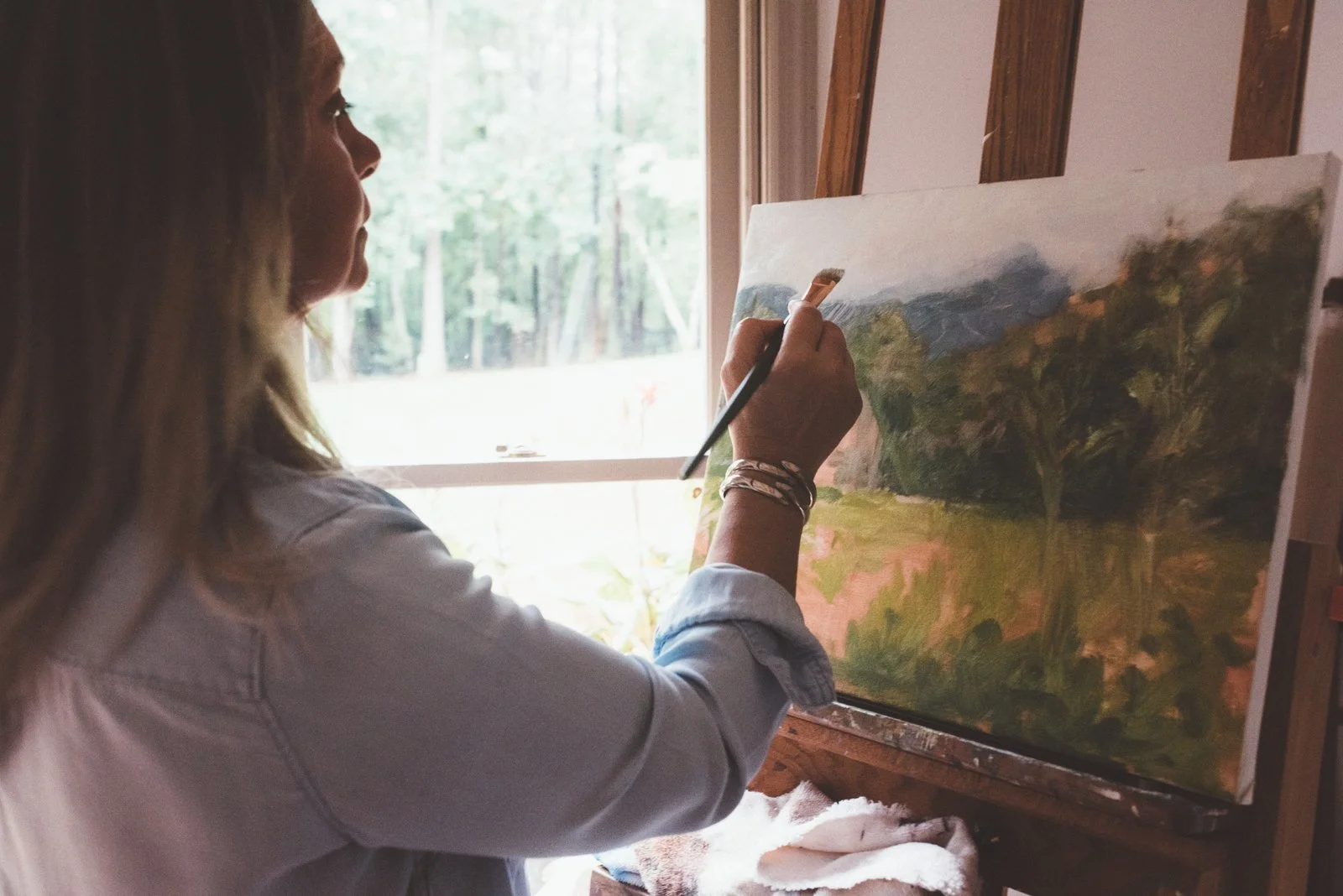 Image of Michaelann Bellerjeau, a woman painting a landscape on a canvas in a room with a window showing trees outside.