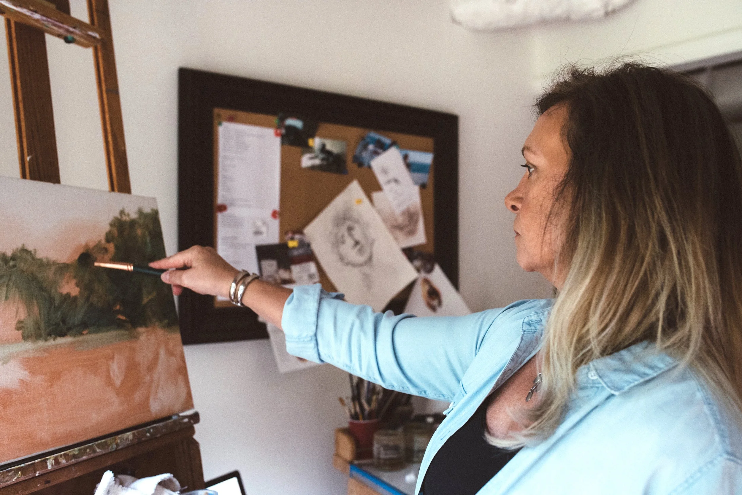 Image of Michaelann Bellerjeau, a woman painting with a brush on a canvas in her studio, pointing to her artwork depicting a landscape with trees and a pink sky.