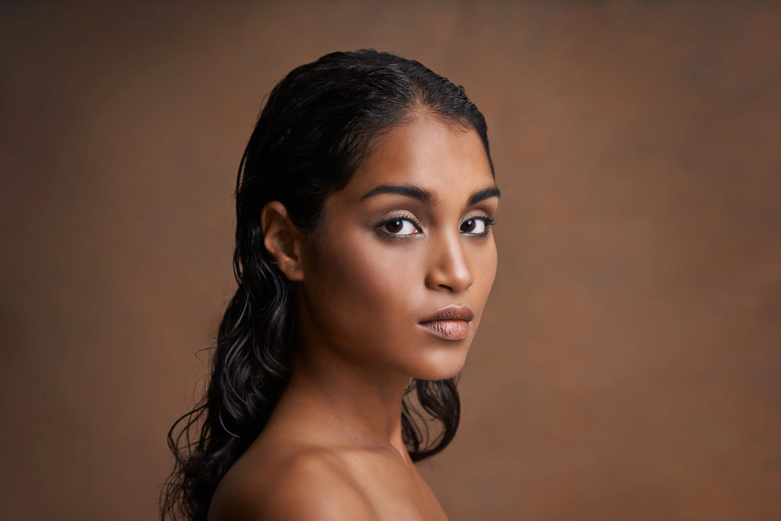 A portrait of a young woman with dark wavy hair, medium skin tone, and makeup, looking at the camera against a brown background.