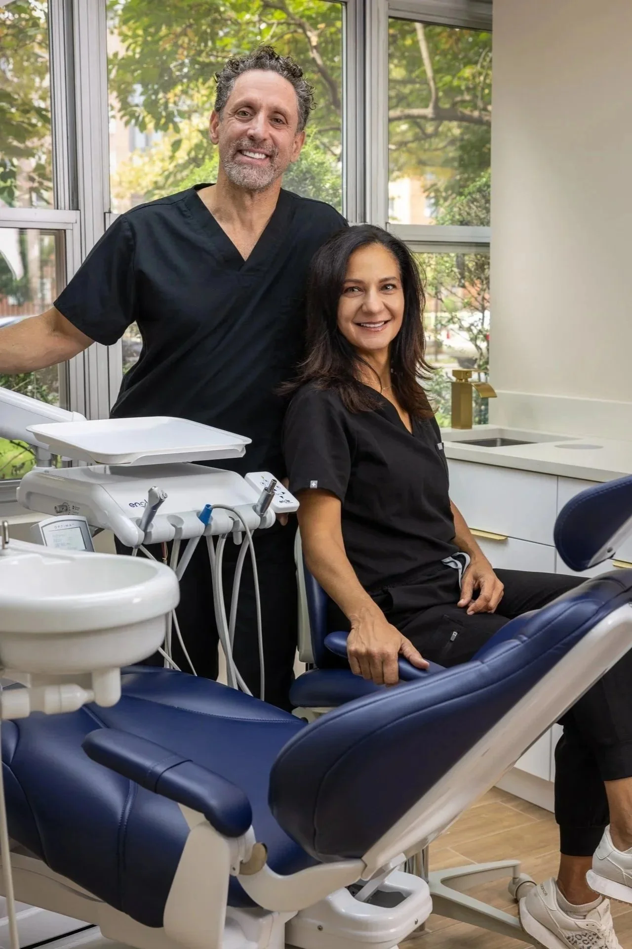 A male dentist with curly hair and a beard stands next to a seated female patient with shoulder-length dark hair in a dental office. The patient is smiling, wearing black scrubs, and sitting in a dental chair with dental equipment nearby. The office has large windows with a view of trees outside.