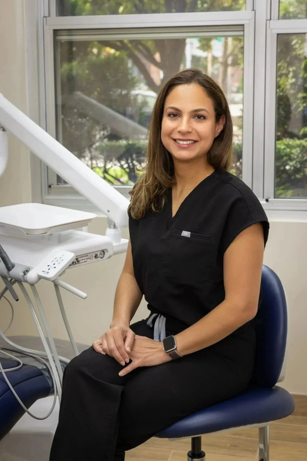 A woman sitting in a dental office, smiling at the camera, wearing black scrubs and a smartwatch, with dental equipment and a window in the background.