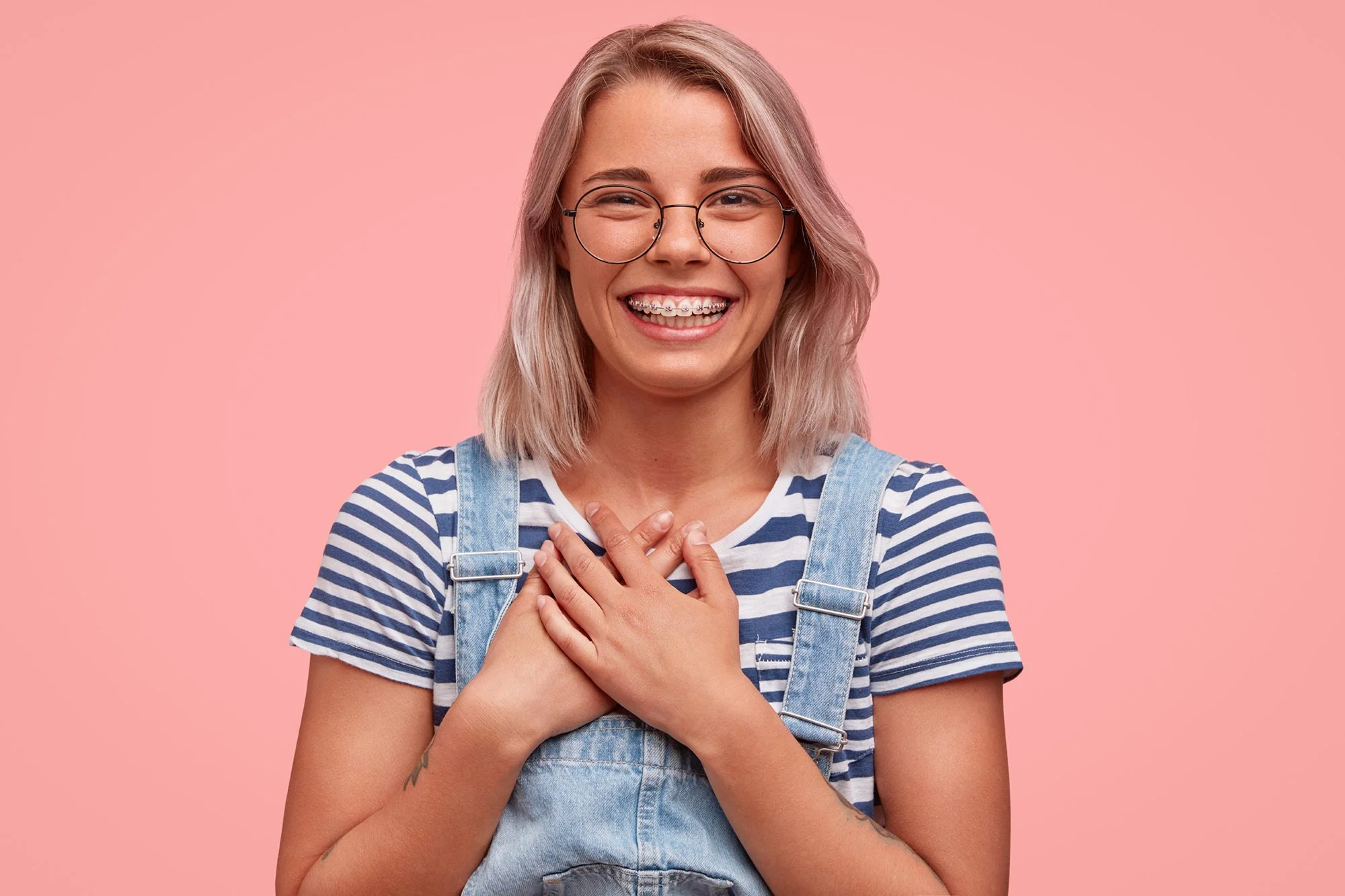 A young woman with blonde hair, wearing round glasses, a striped t-shirt, and denim overalls, smiling and placing her hands on her chest against a pink background.