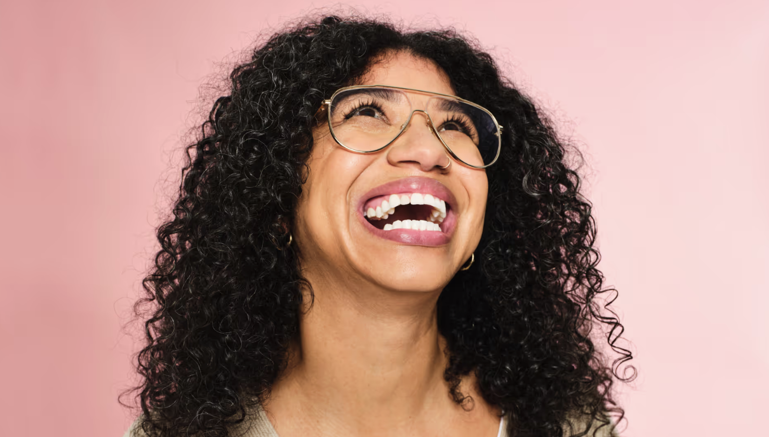 A woman with curly black hair and glasses laughing against a pink background.