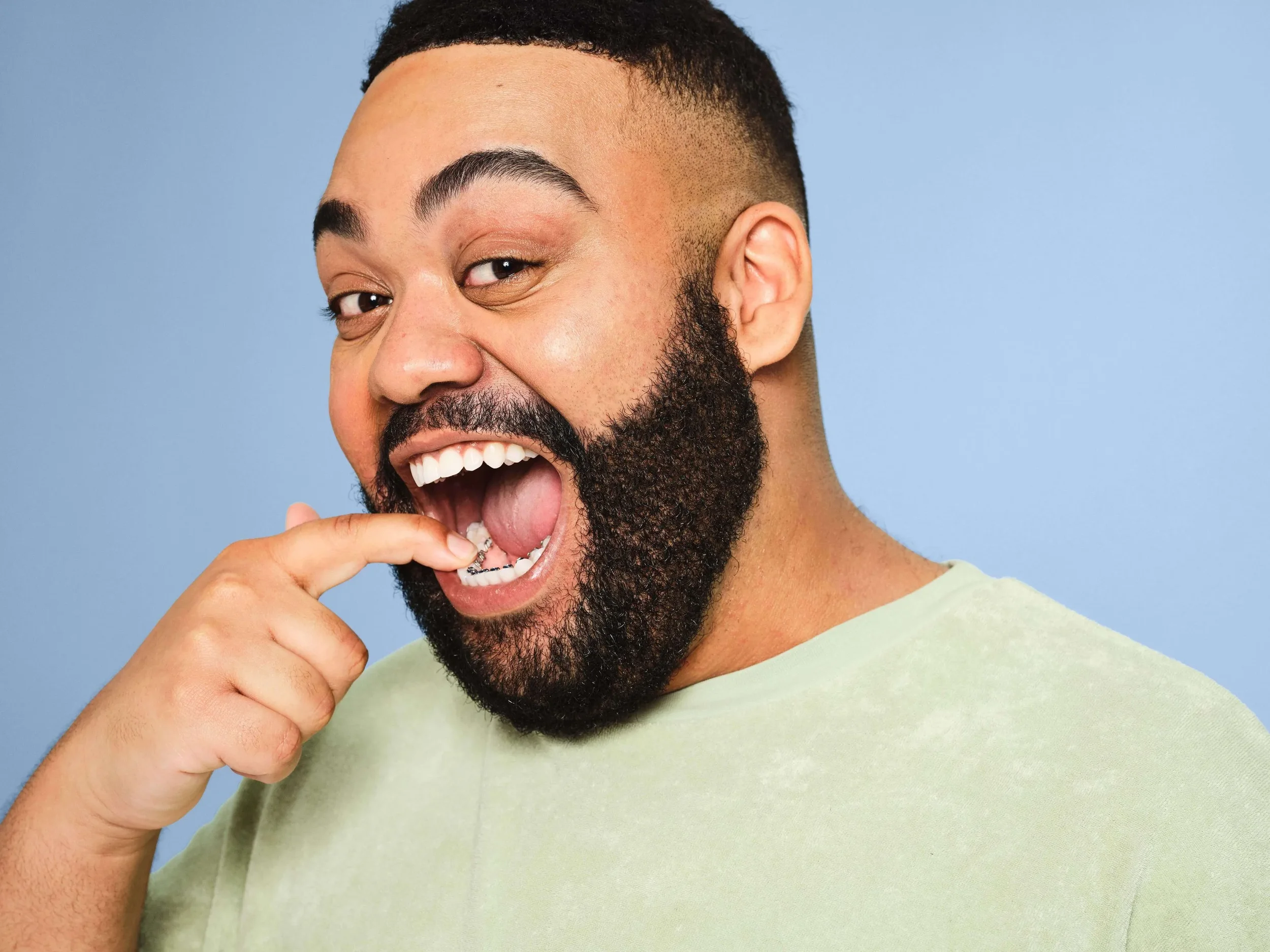 A man with a beard and mustache, smiling broadly, wearing a light green T-shirt, with a blue background.